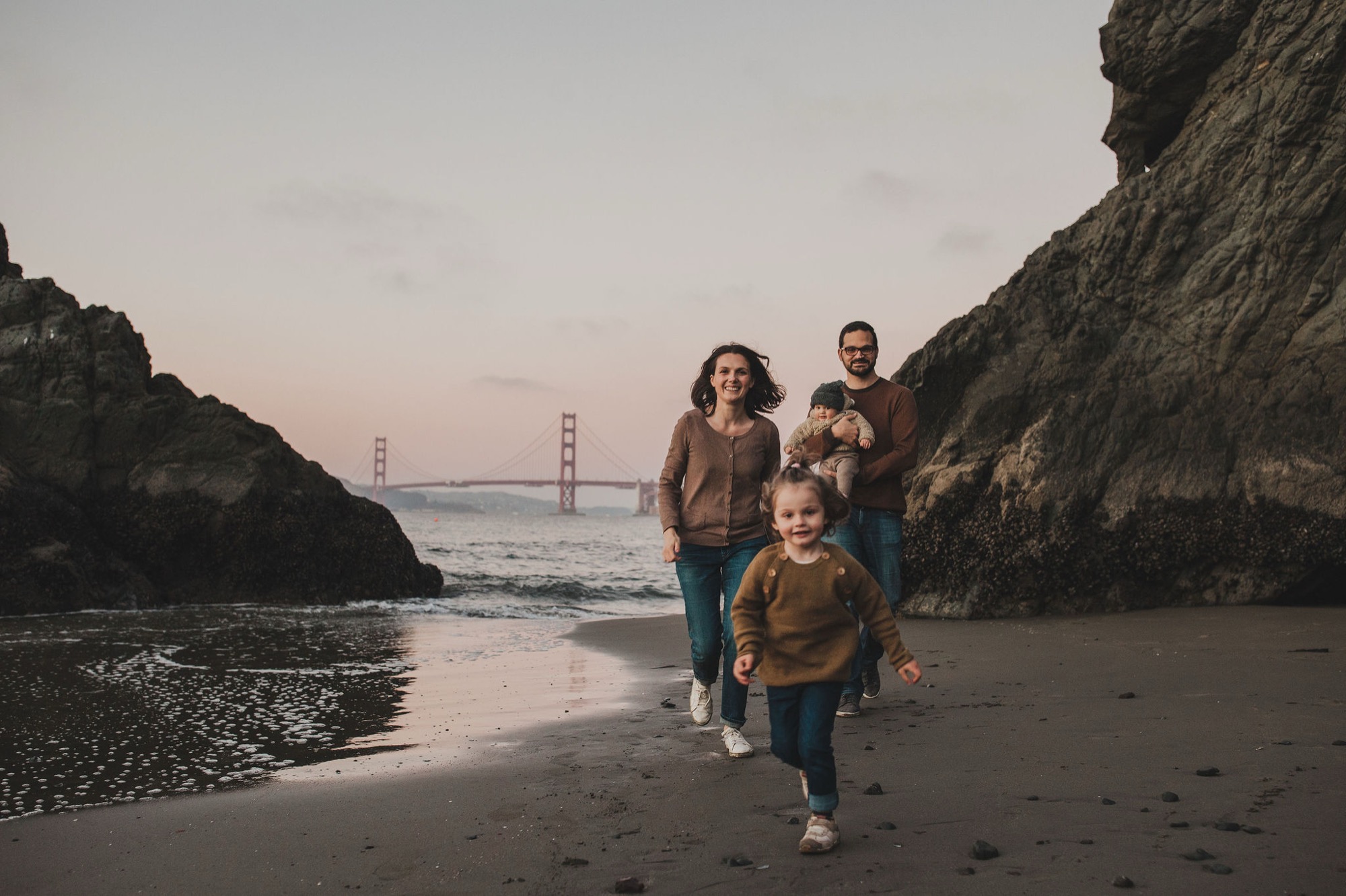 A family of four walks toward the camera on wet sand between towering rock formations, the Golden Gate Bridge glowing softly in the dusky background