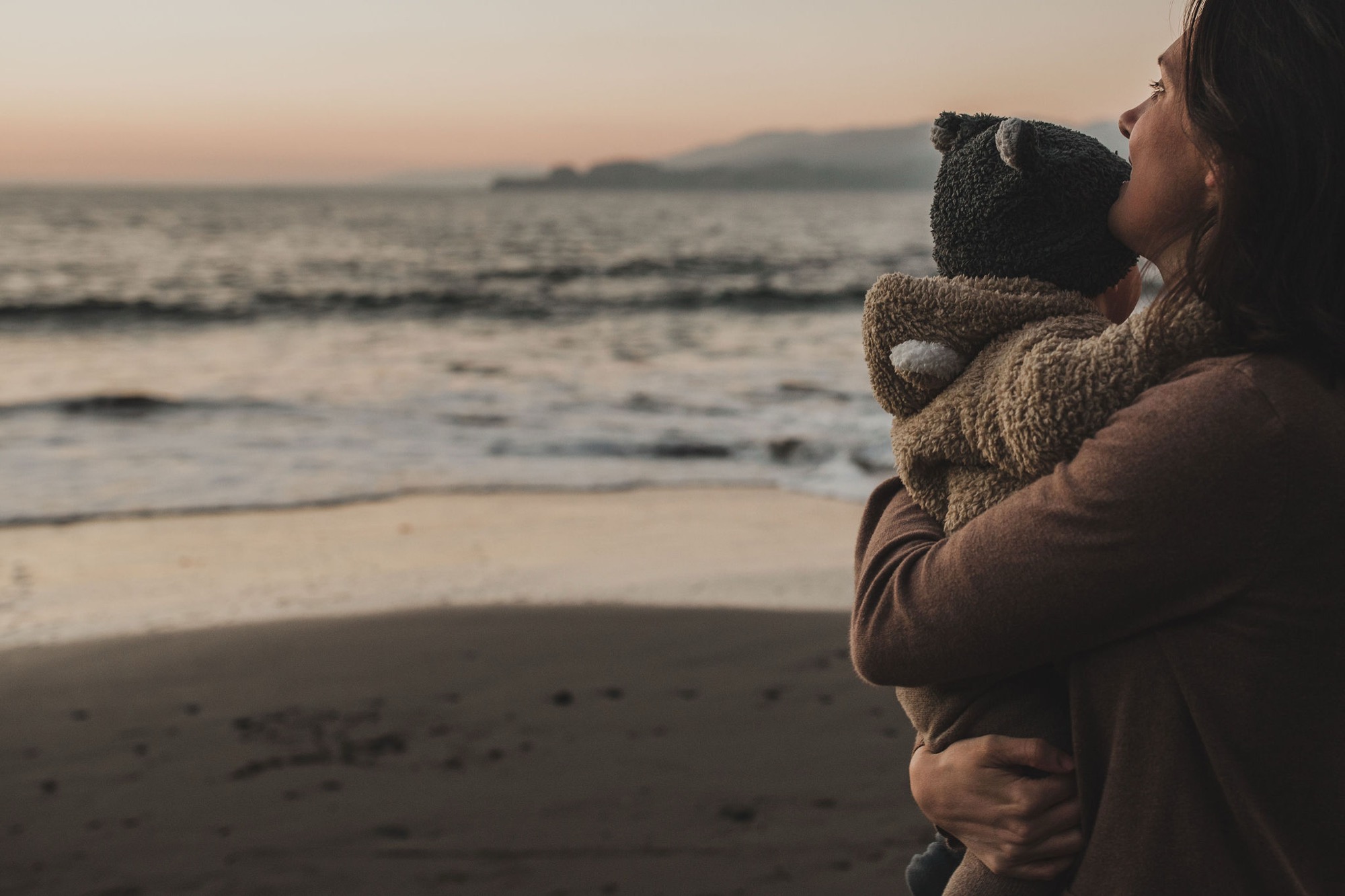 A woman holds her child from behind on the beach at golden hour, both gazing toward the misty horizon and distant shoreline