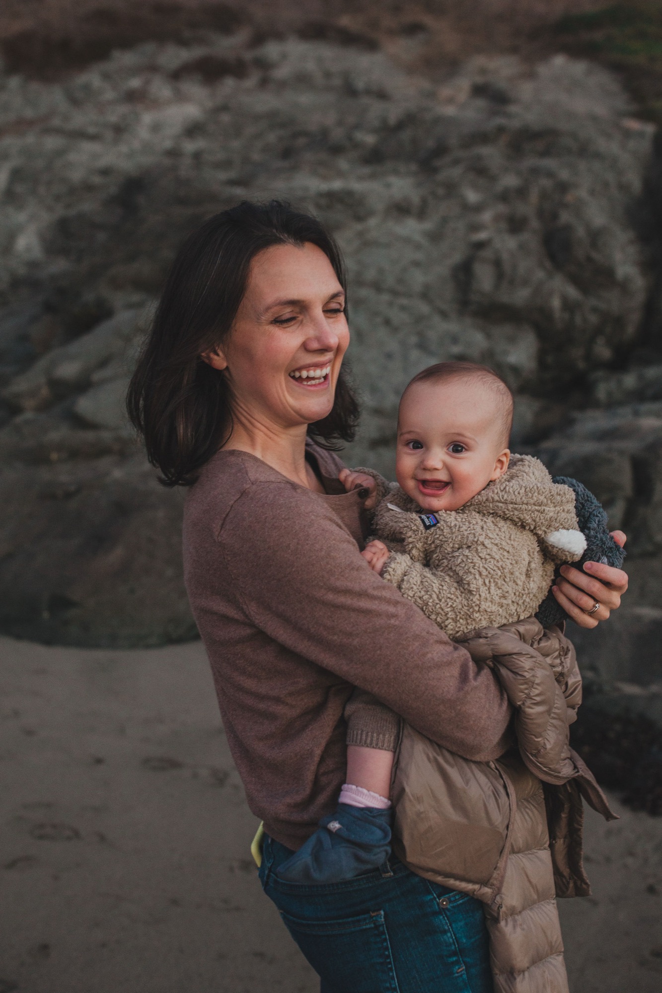 Woman holds smiling toddler wrapped in a tan blanket against weathered gray rock formations, both content in soft natural light
