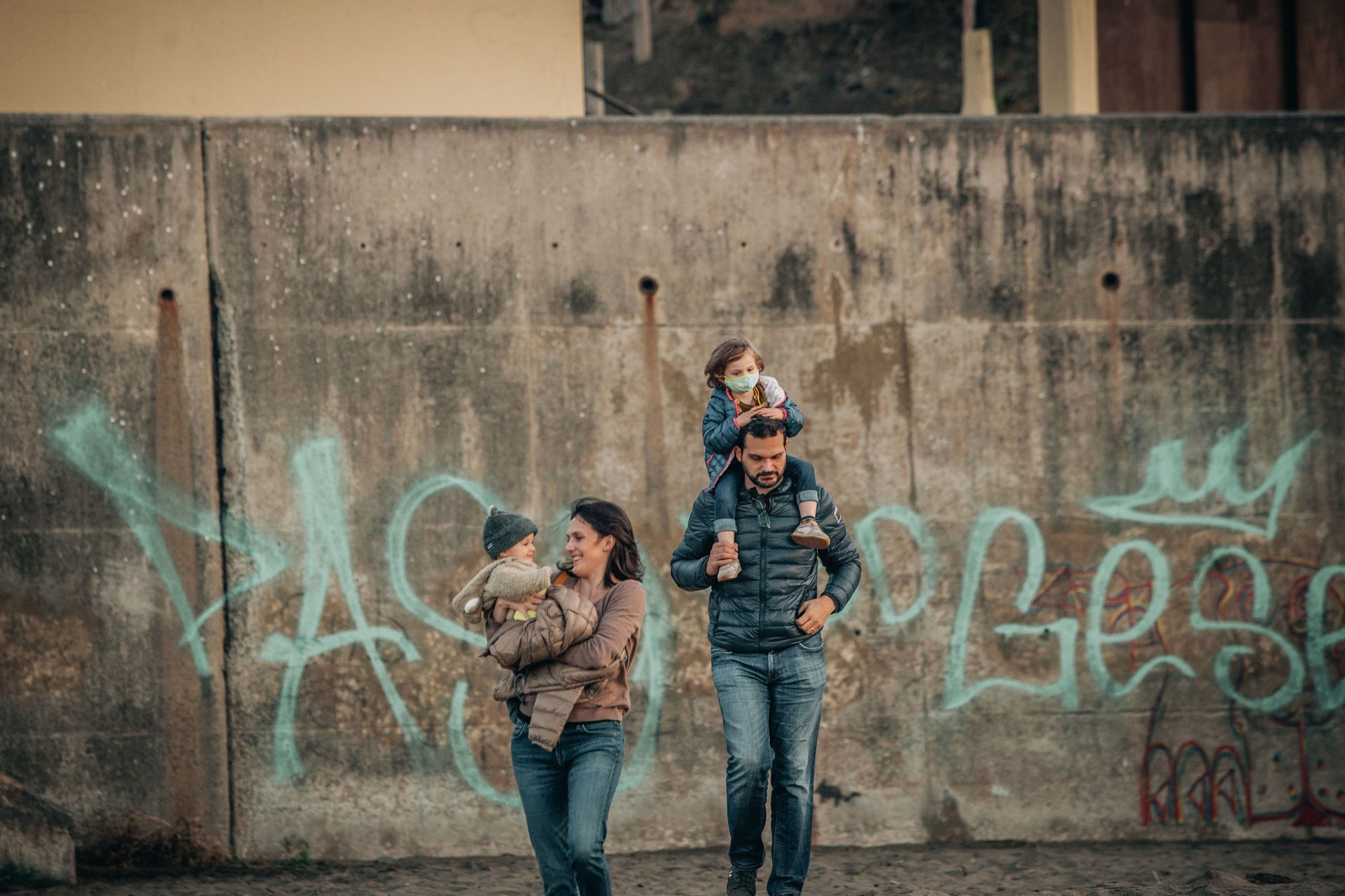 Two children laugh and run toward the camera past a weathered concrete wall tagged with turquoise graffiti