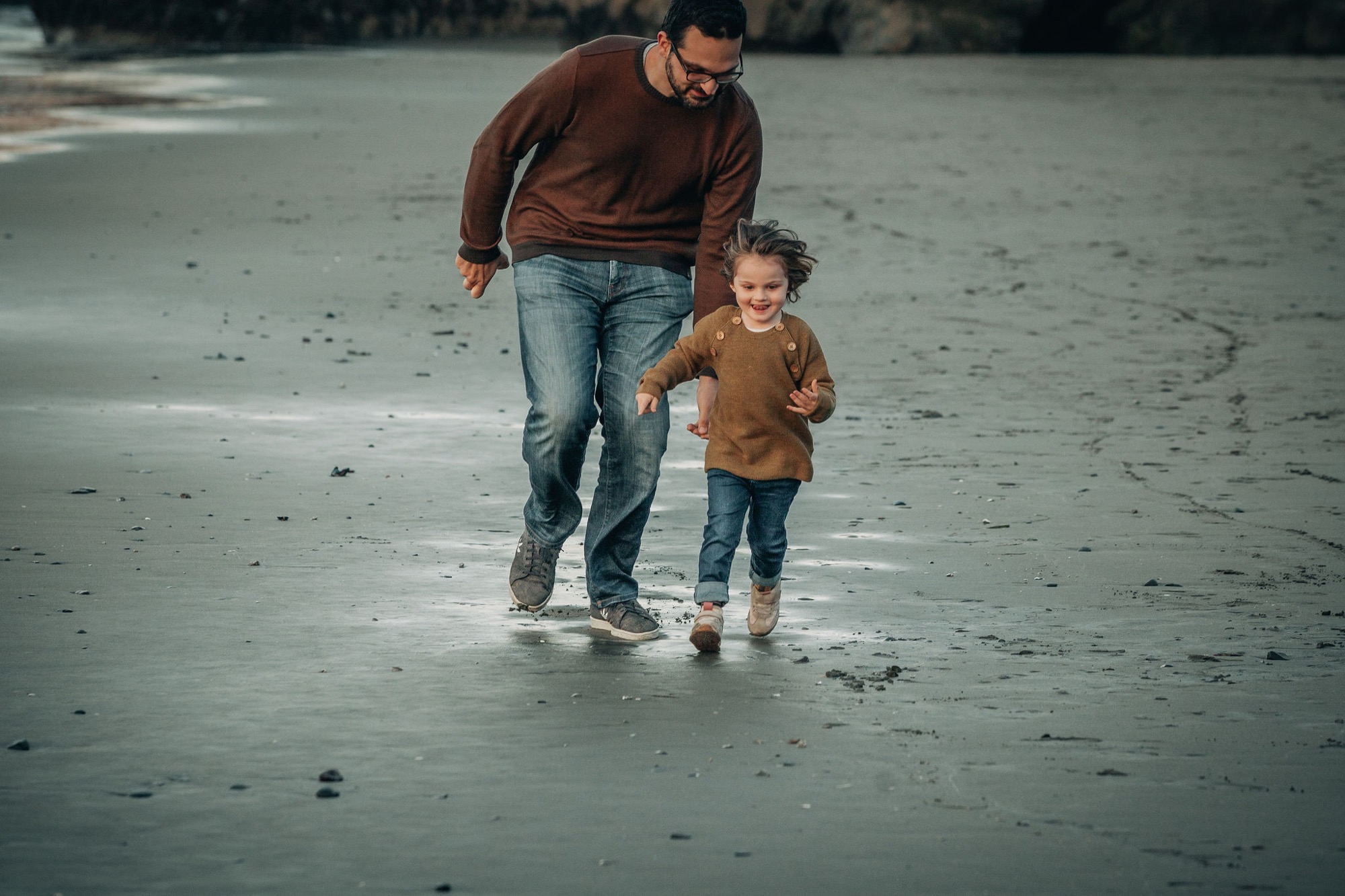 A father and young child run together across wet sand at the beach, their figures casting shadows in the golden afternoon light