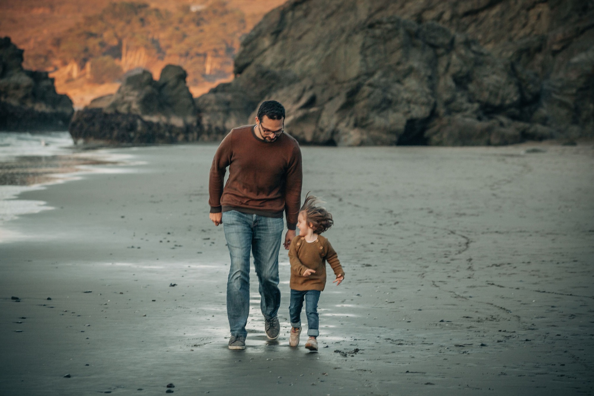 A father and young child walk hand-in-hand across wet sand as golden evening light illuminates towering coastal cliffs behind them