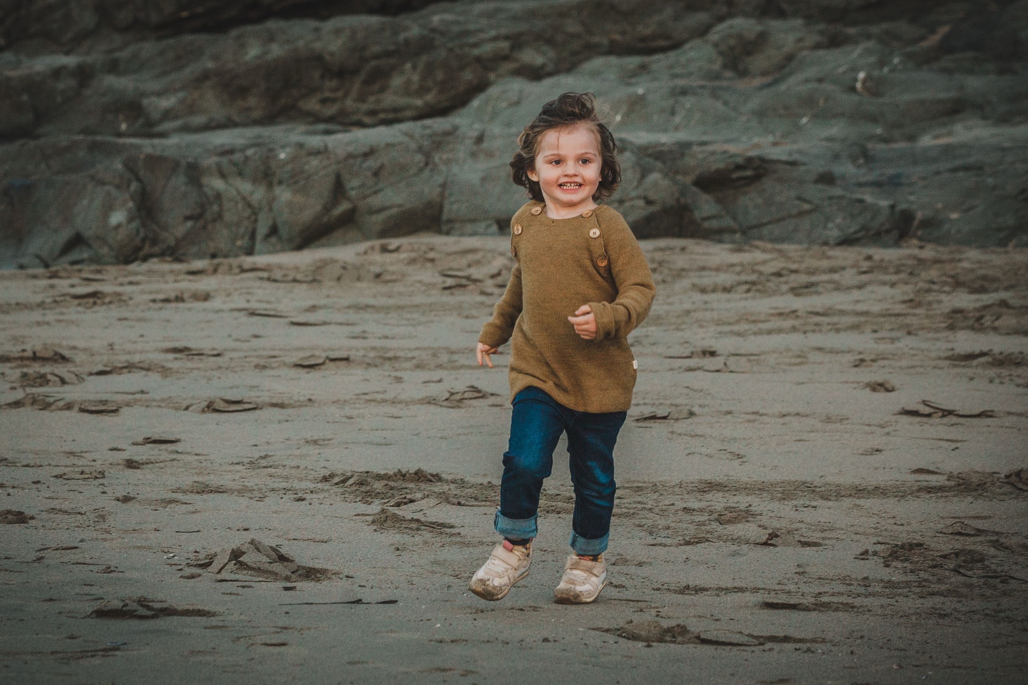 A young child in a mustard sweater and blue jeans smiles broadly while running across wet sand at the beach, rocky cliffs looming behind