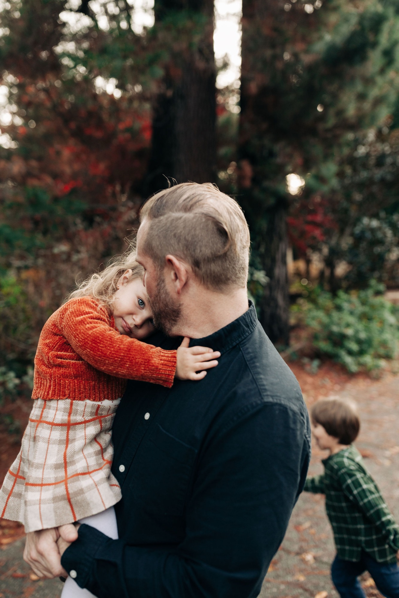 A father holds his young daughter on his shoulders while a second child stands nearby, backlit by golden afternoon light filtering through autumn trees