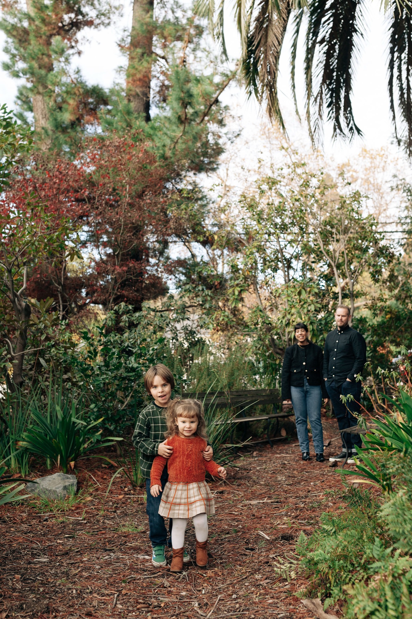 A young child in a red dress leads the way down a shaded woodland path while parents follow behind through dappled garden light