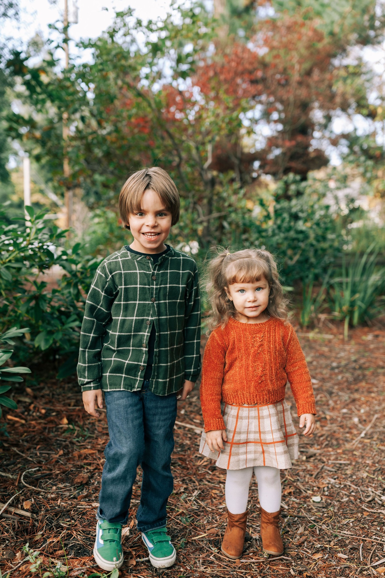 Two young children stand together on a garden path, the older boy in a green plaid shirt smiling warmly while the younger girl in a rust-colored knit sweater gazes downward