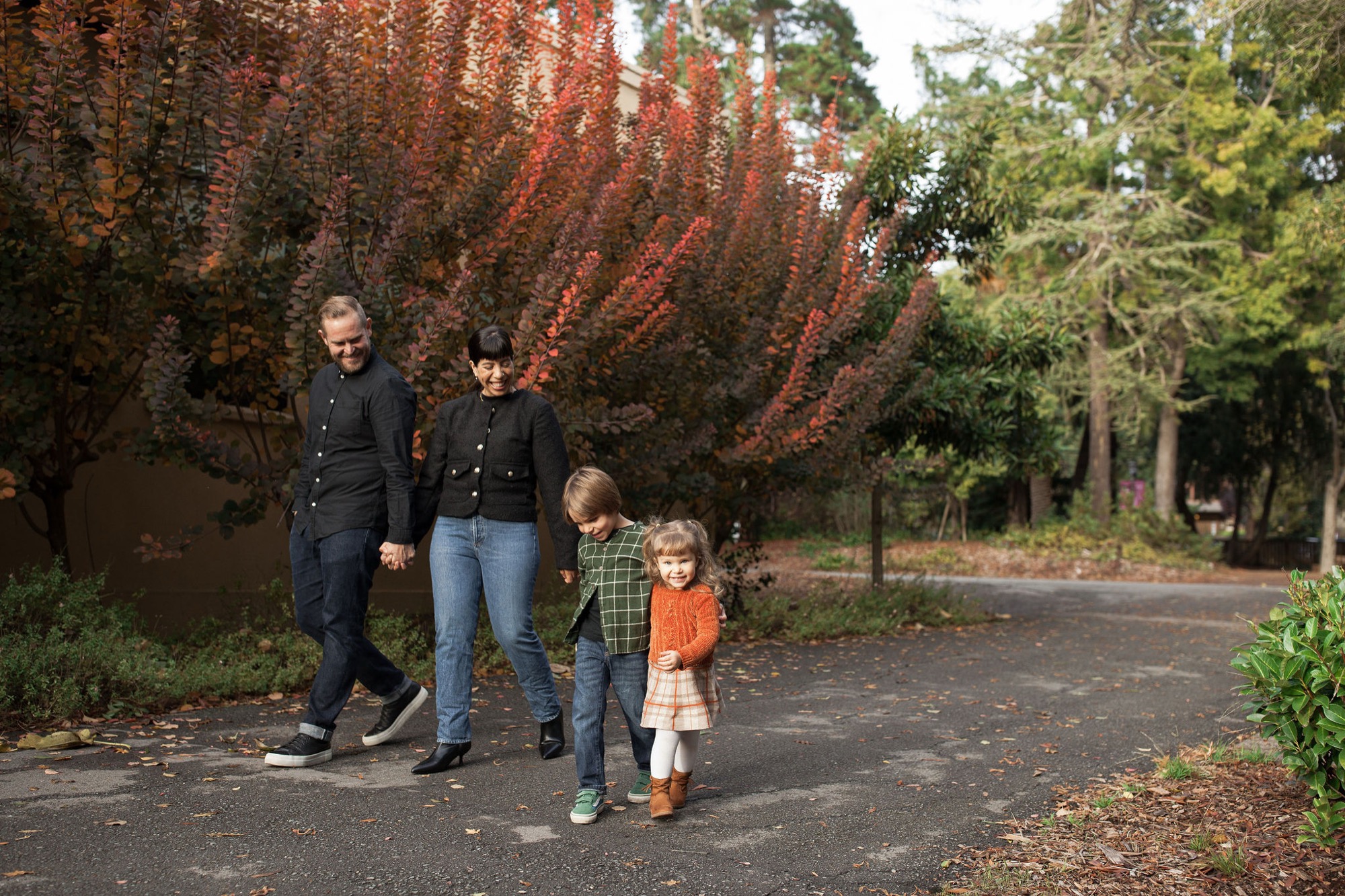Family of four walks together on a tree-lined park path, with autumn foliage glowing red-orange behind them