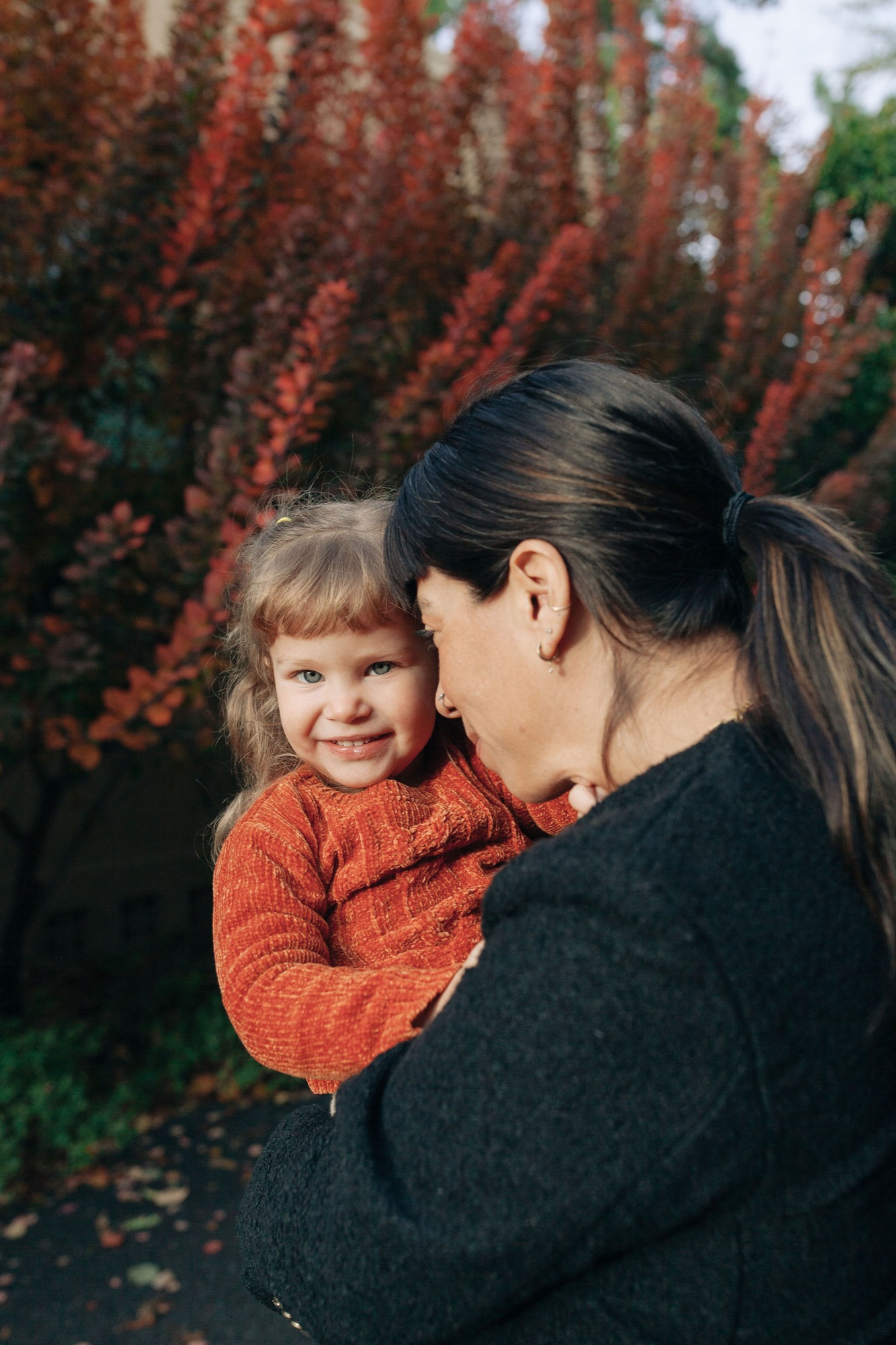 A woman holds her smiling toddler in an orange sweater against a backdrop of deep red ornamental grasses