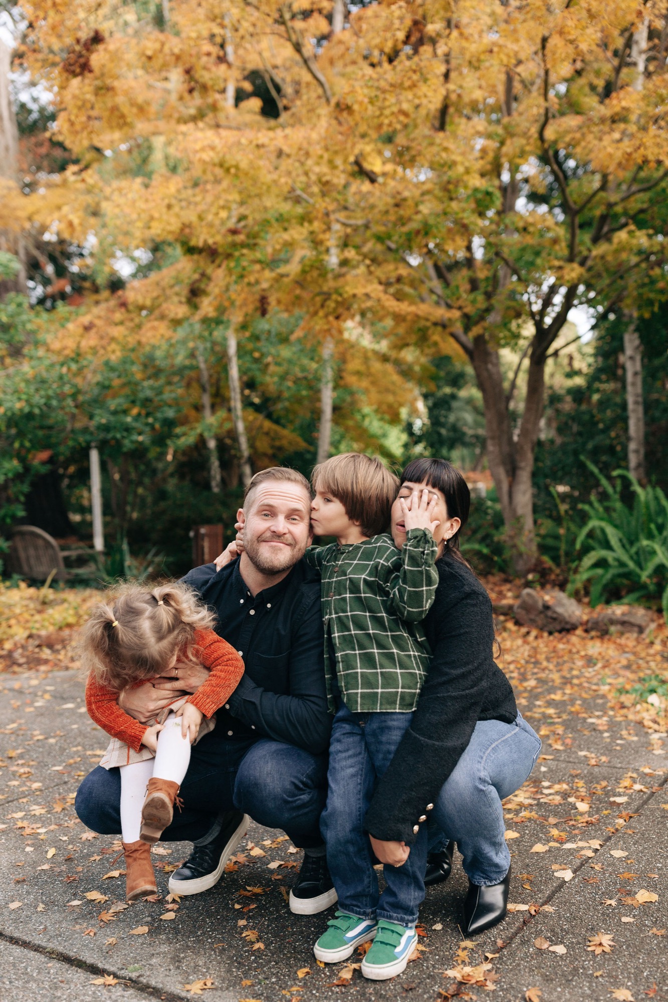 Father crouches with two young children on an autumn path, all smiling warmly while golden leaves frame the scene behind them