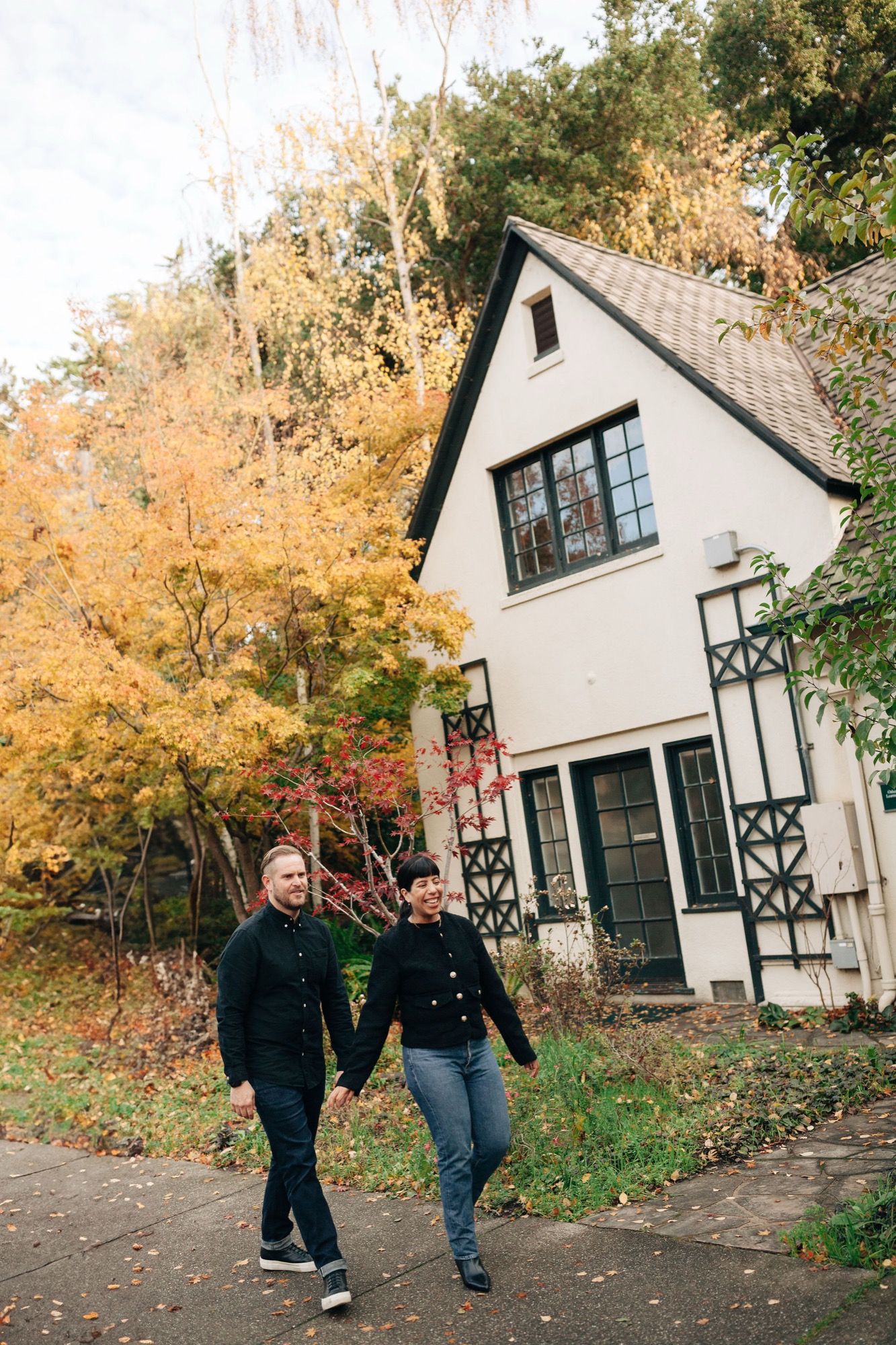 Two people jog together on a tree-lined path beside a charming Tudor-style house on an autumn afternoon