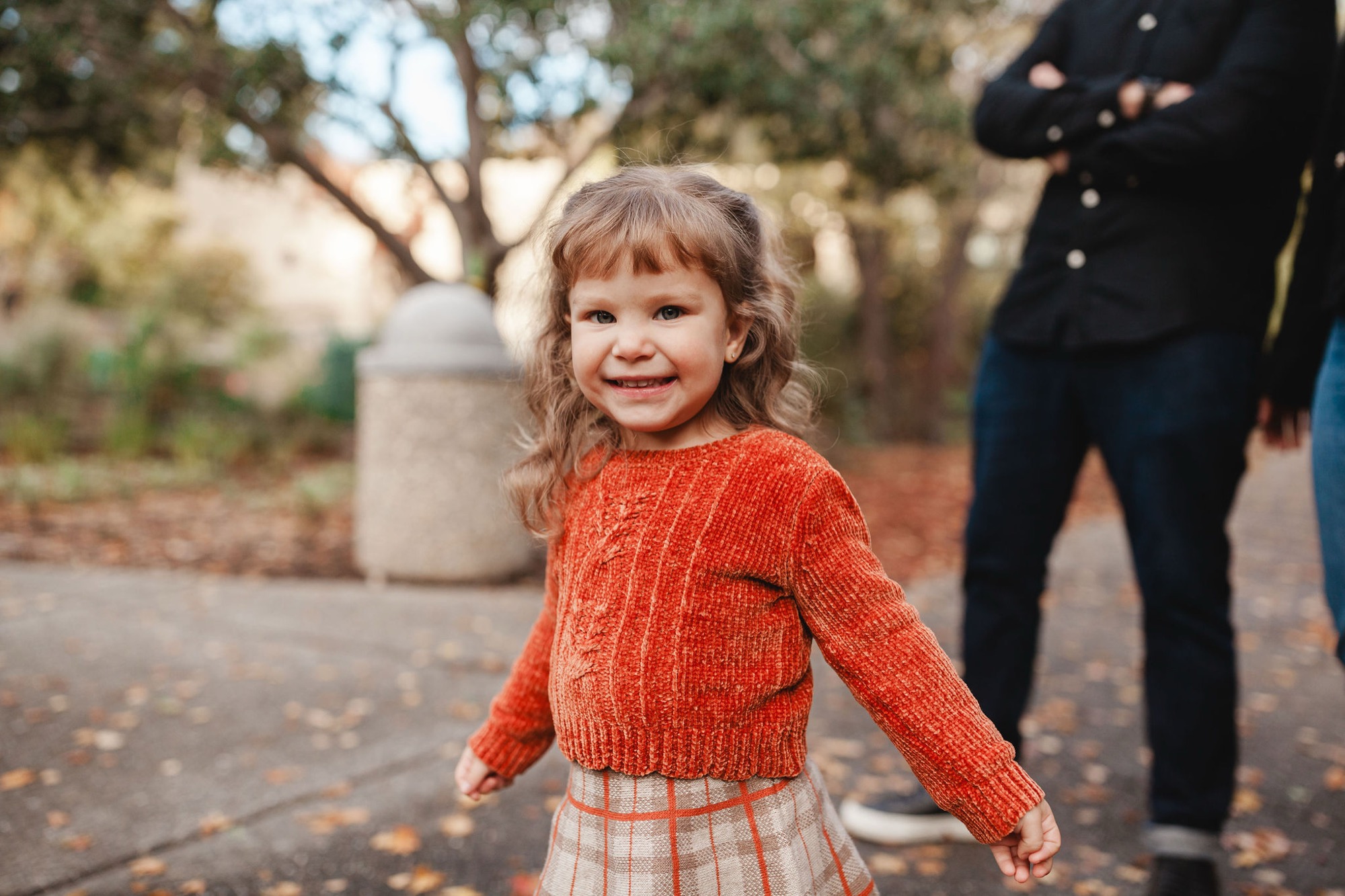 A young girl in a rust-colored ribbed sweater smiles warmly at the camera while standing on a paved path, with blurred family members visible behind her