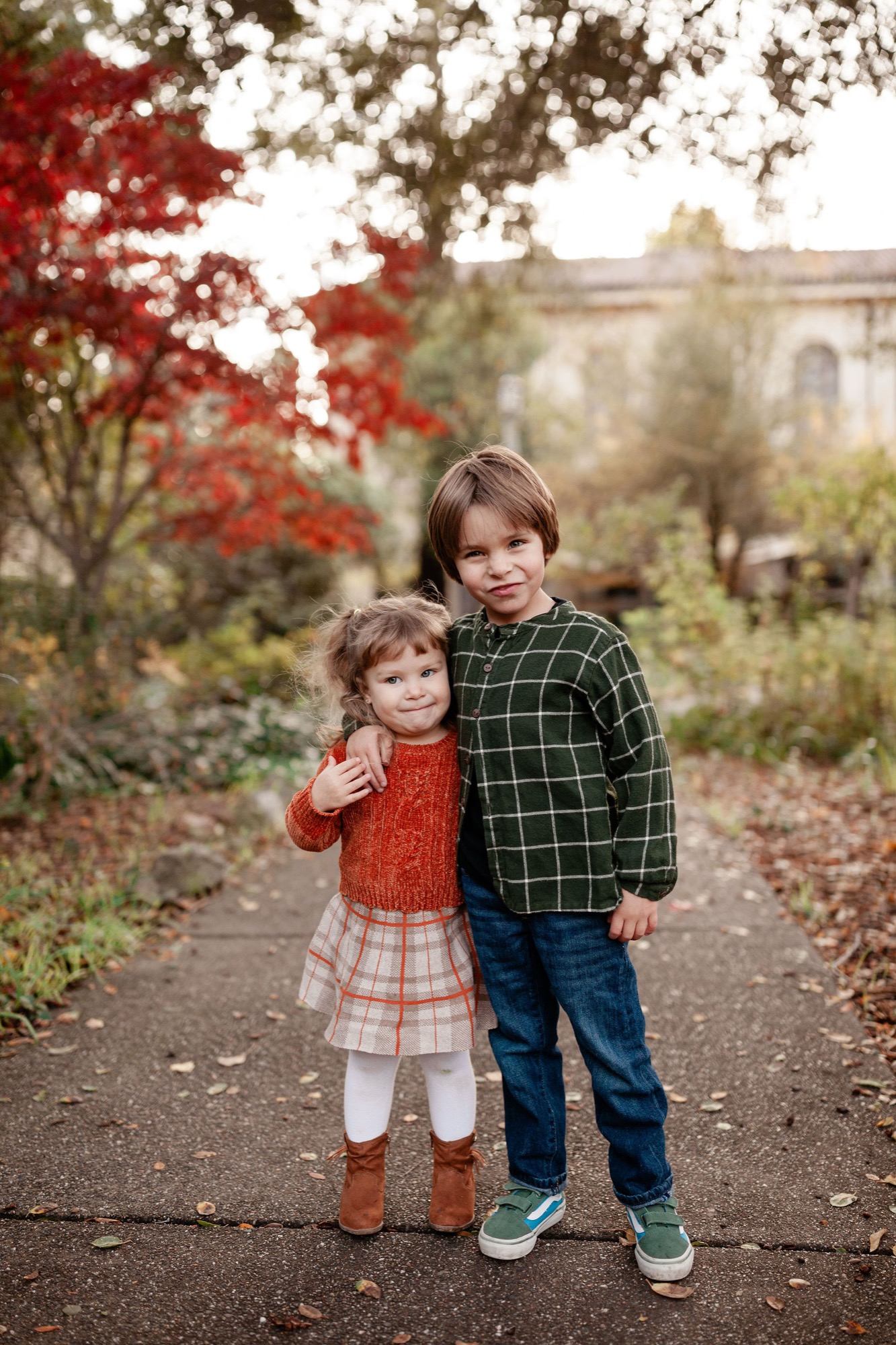 Two young children stand arm-in-arm on a garden path, autumn leaves glowing red above them in soft afternoon light