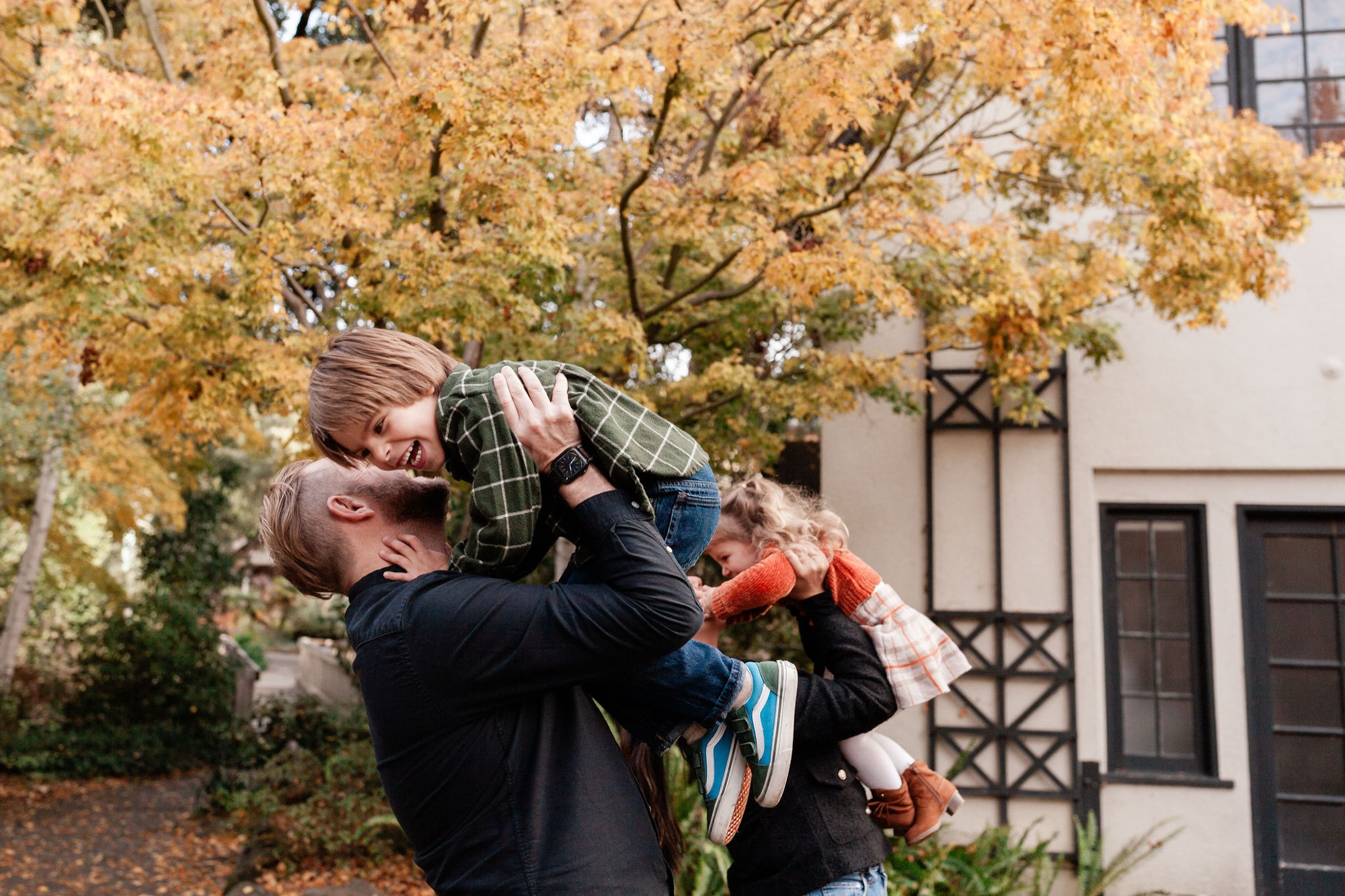 Father lifts laughing child overhead against golden autumn foliage and residential building backdrop