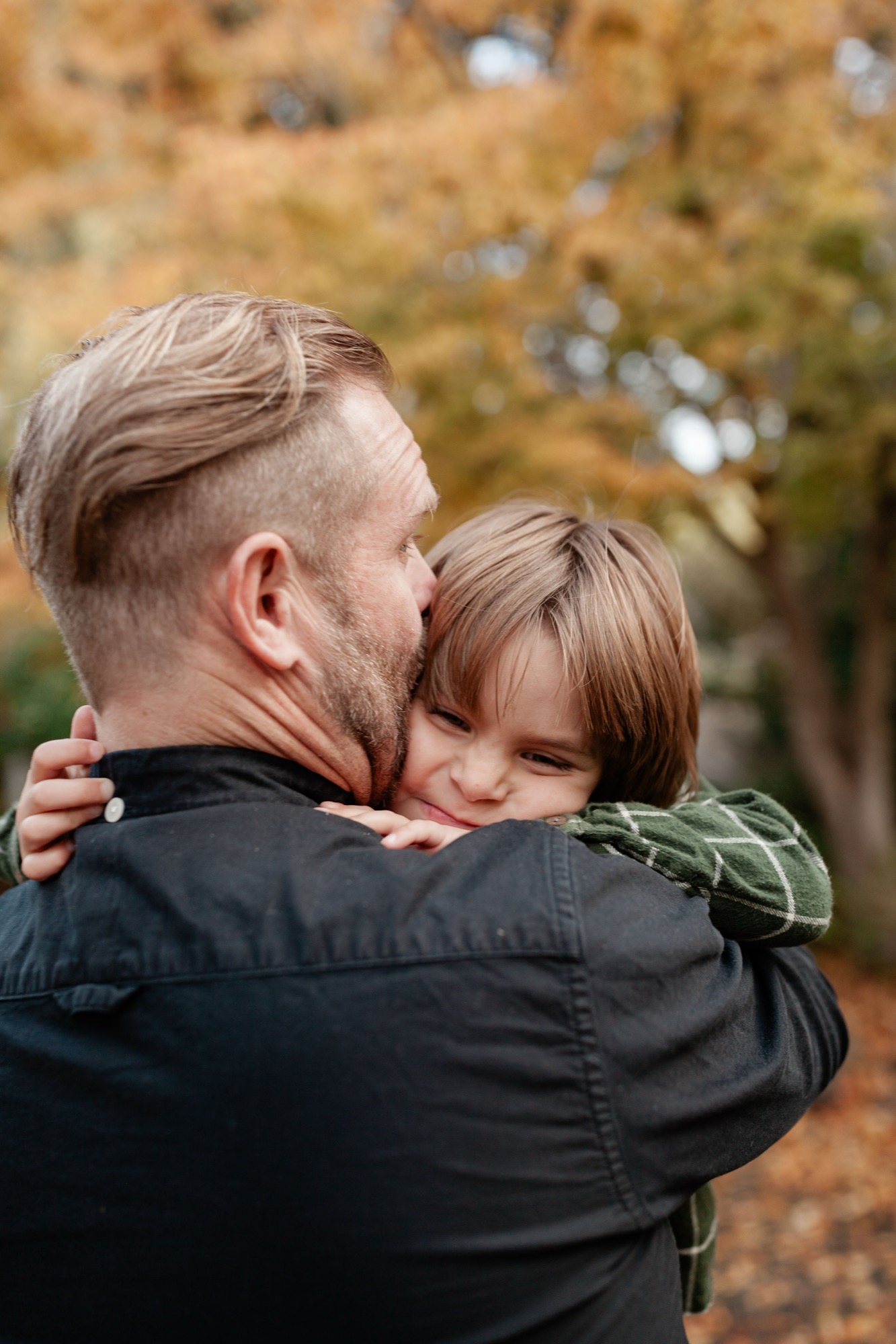 A father carries his young daughter on his back in autumn woods, both wearing earth-toned clothing against golden fall foliage