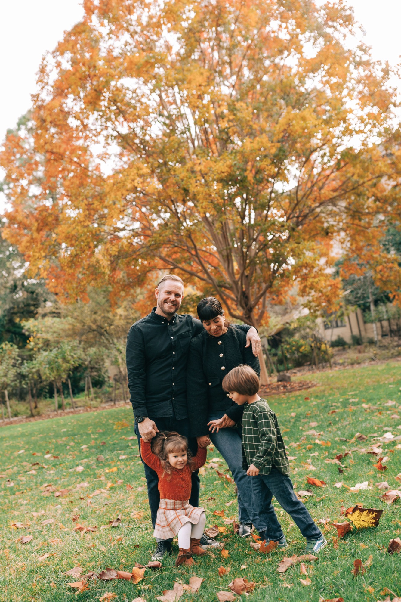 Father stands with three young children beneath a golden autumn tree, their bodies relaxed and close together on a grassy slope