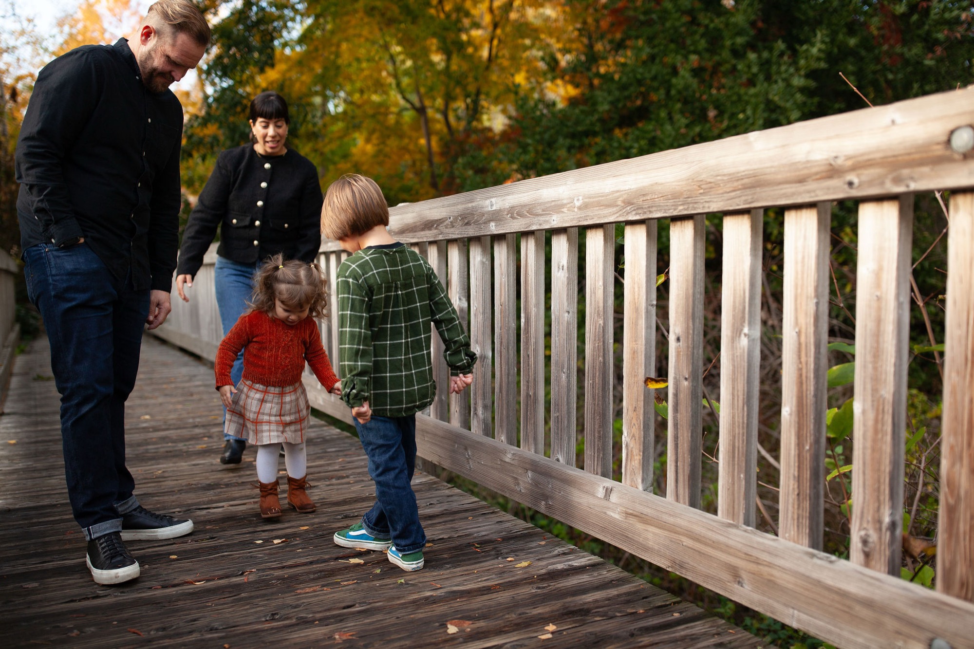 Adults and children walk together on a wooden bridge deck, autumn foliage glowing behind them in warm golden light