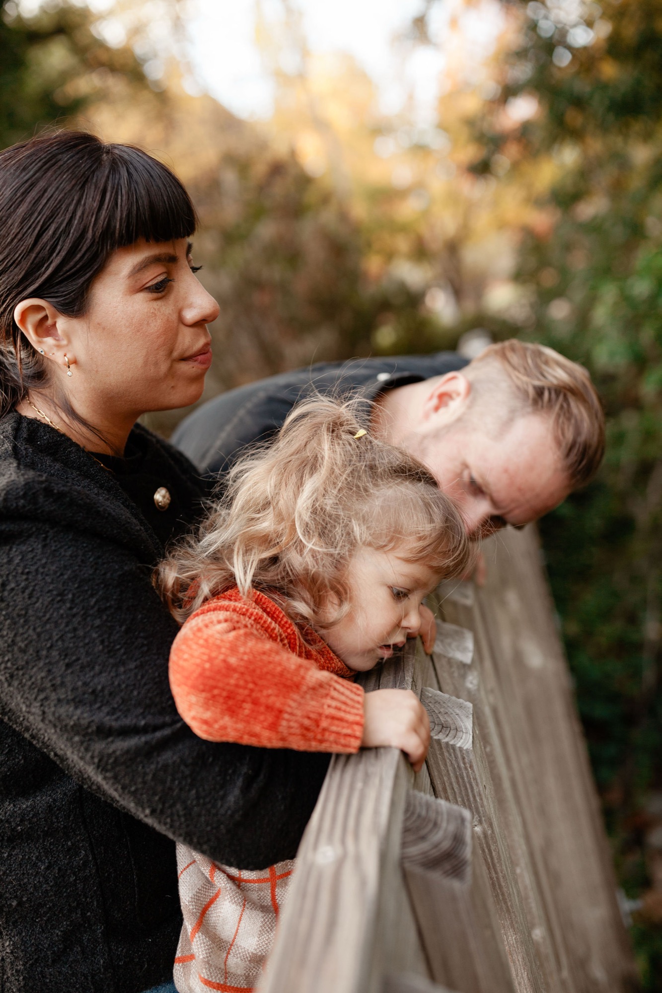Woman holds two young children against her chest in an autumn woodland setting, their faces relaxed and intimate in soft golden light