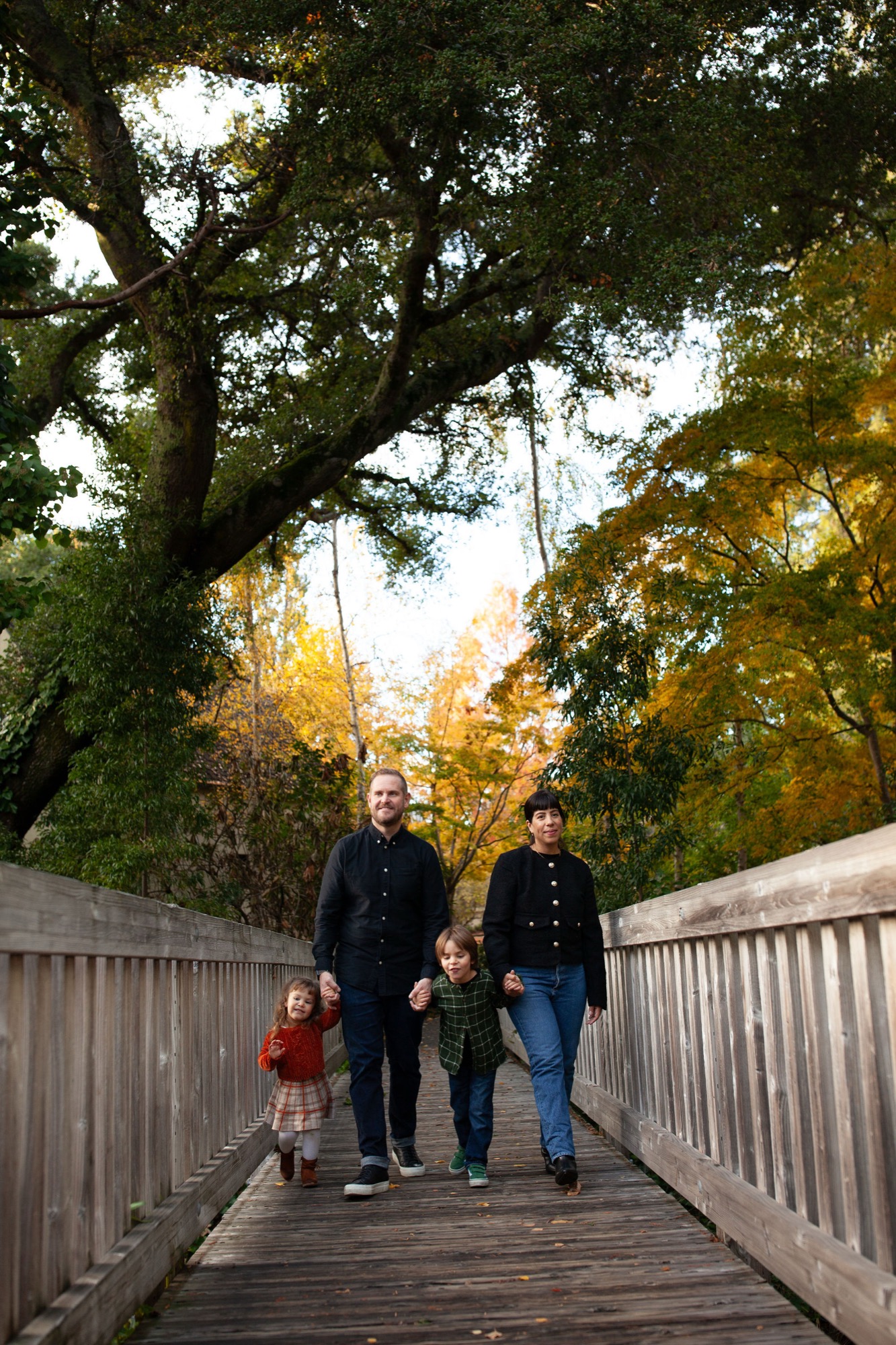 Family of four walks across a wooden bridge toward golden autumn trees, bathed in warm afternoon light filtering through the canopy