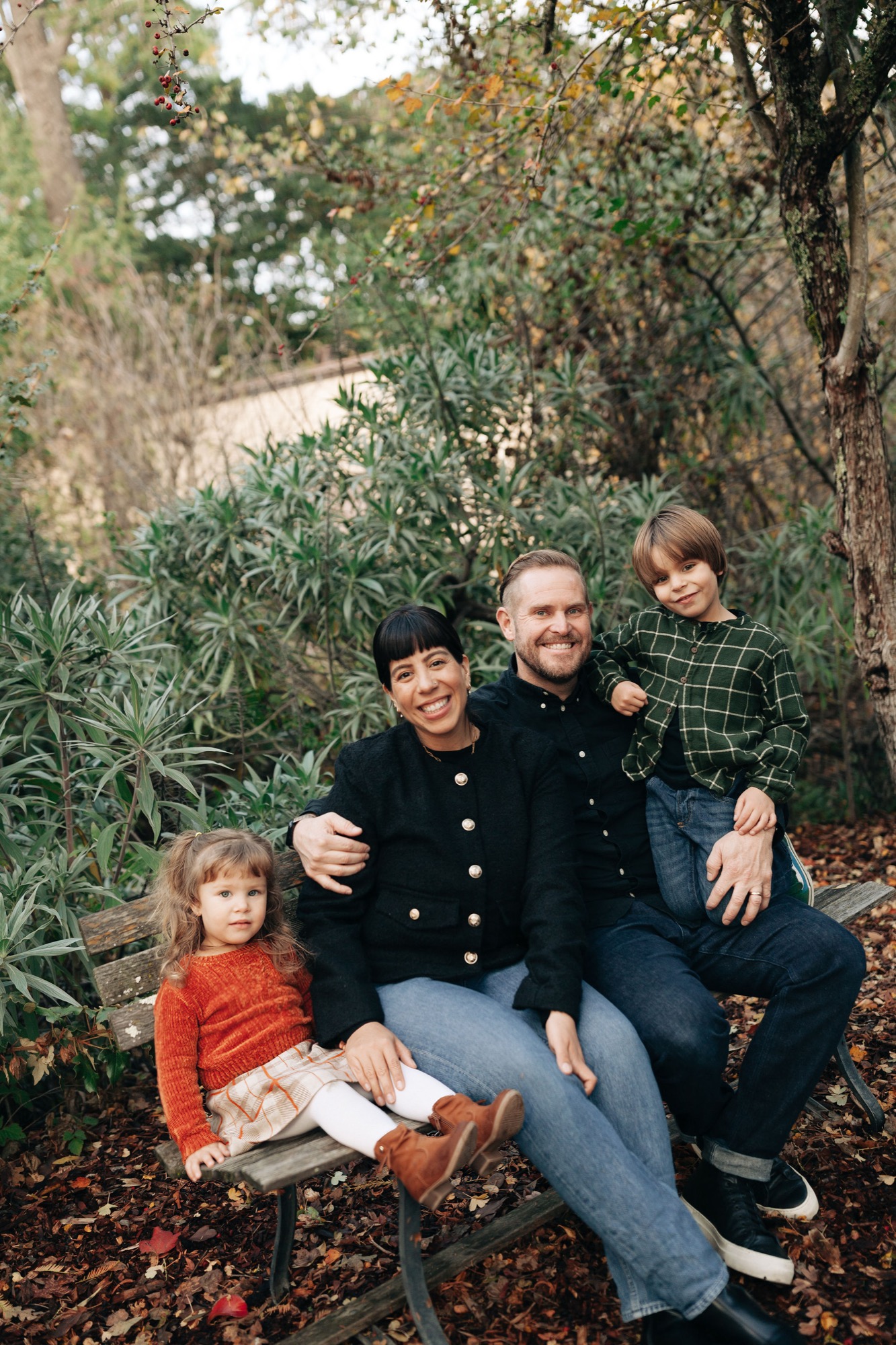 A father sits with three children among autumn leaves and evergreen shrubs, all smiling warmly at the camera in dappled natural light