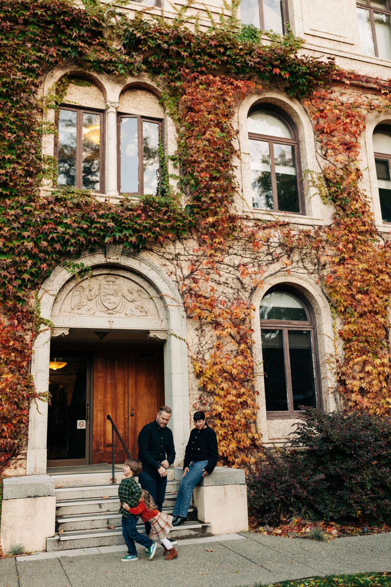 A family of three walks toward a historic ivy-covered brick building entrance on a bright afternoon
