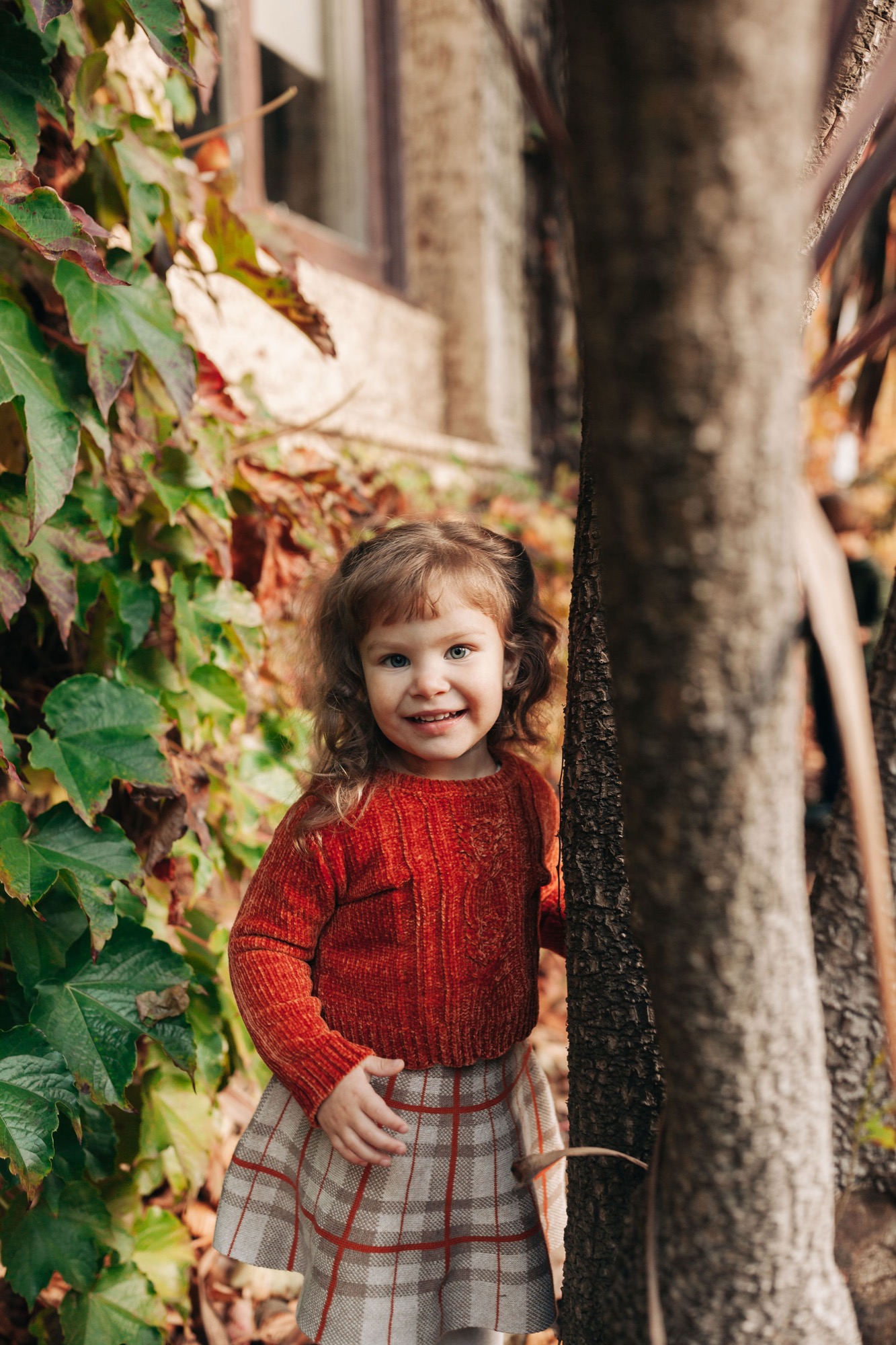 A smiling toddler in a crimson cable-knit sweater and plaid skirt stands between ivy-covered walls and weathered tree trunks