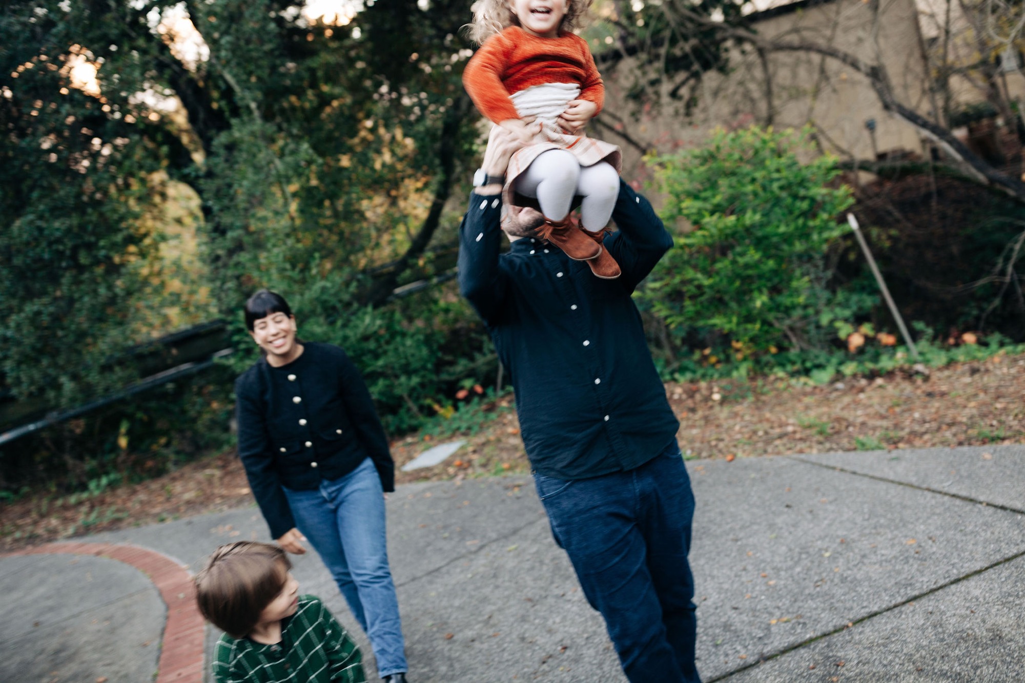 A father tosses his young daughter in the air while two other children watch on a tree-lined residential street