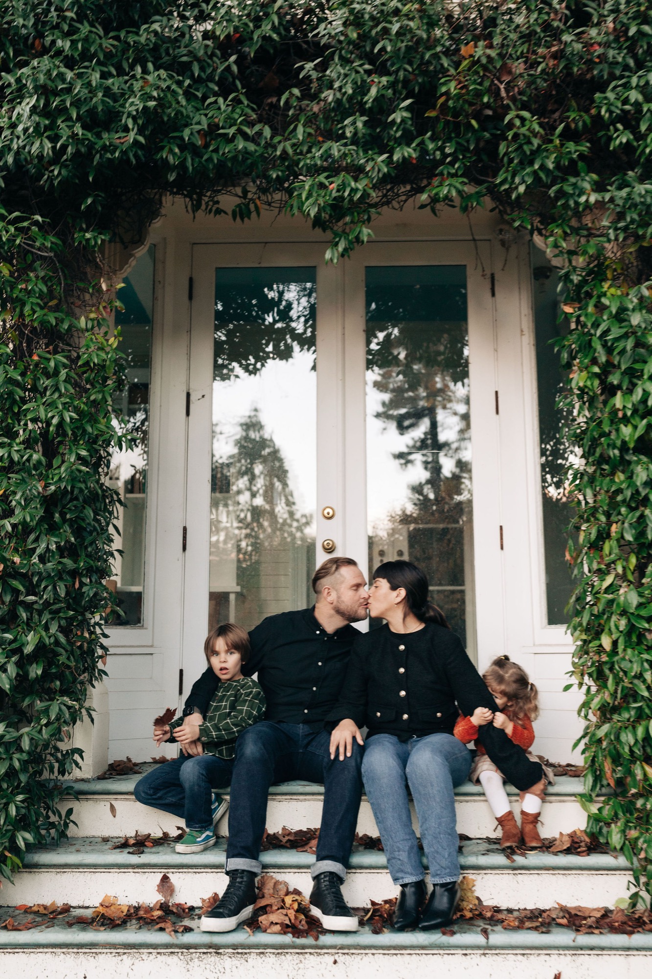 A family of four sits on ivy-framed steps, the parents kissing while children nestle beside them in autumn light