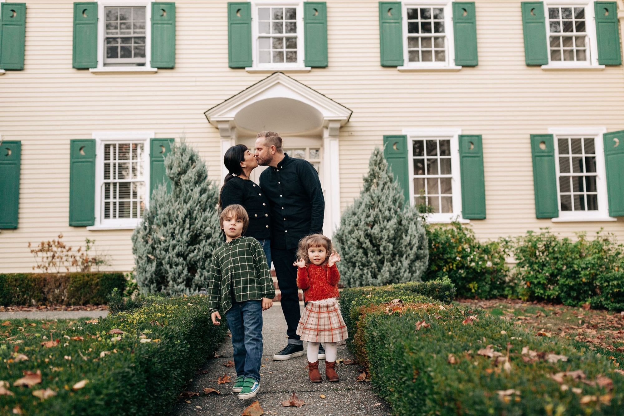 Parents kiss while two young children stand together on a manicured garden path before a colonial home with green shutters