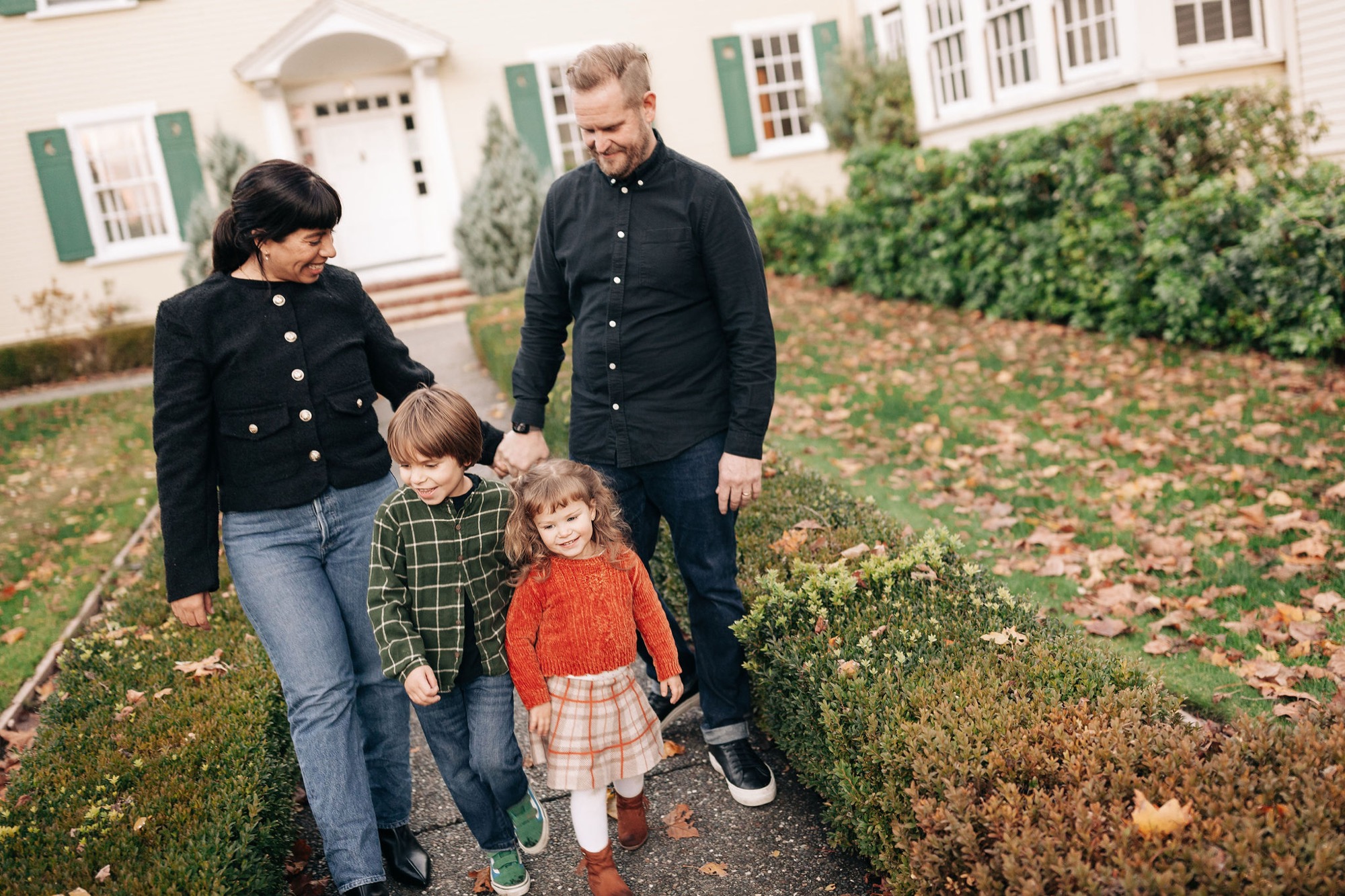 Two adults and two young children walk together down a garden path lined with low shrubs, the brick house visible behind them