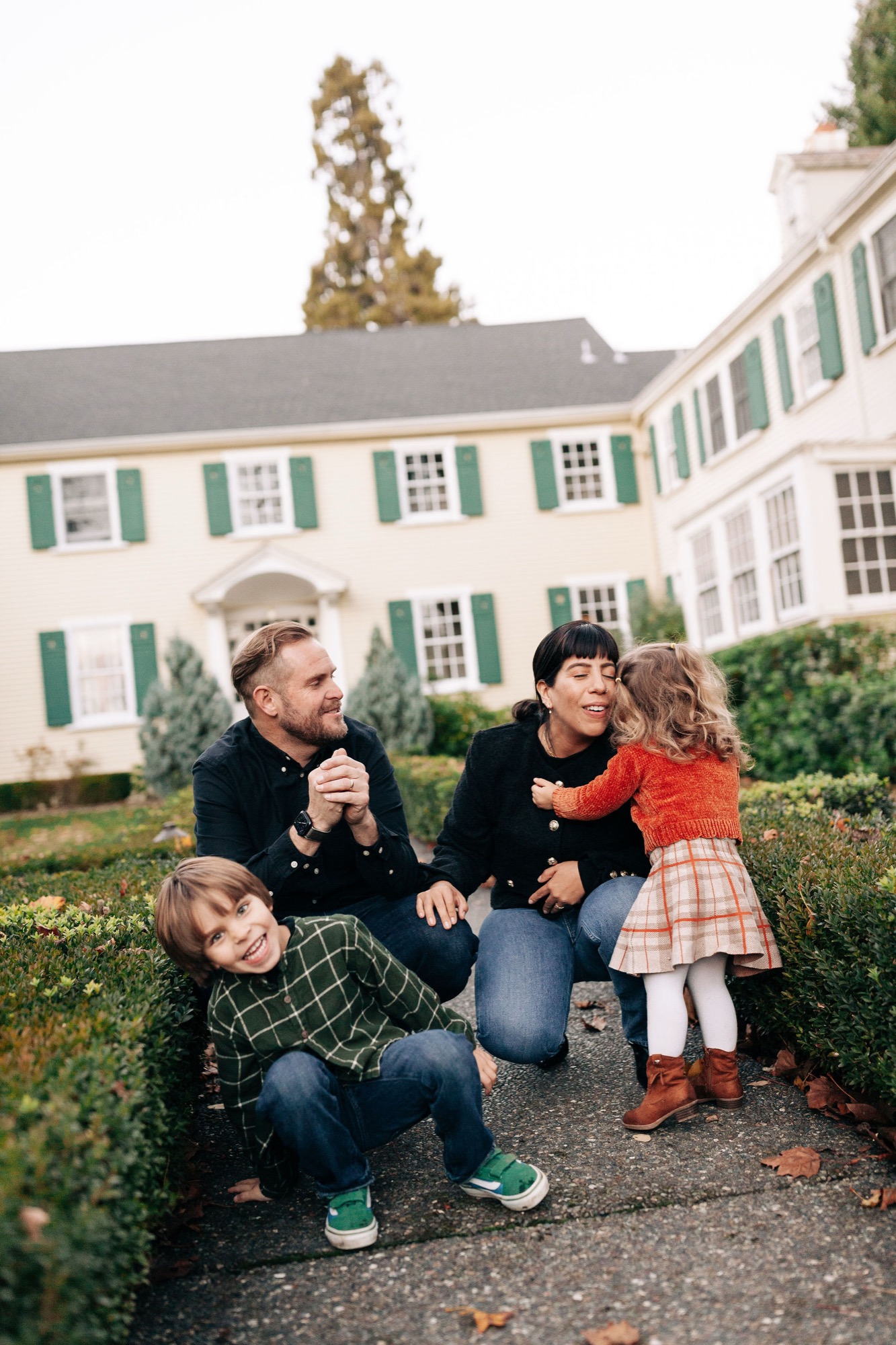Two adults crouch beside two young children on a garden path, all gazing toward the camera with soft afternoon light filtering across the manicured hedges behind them