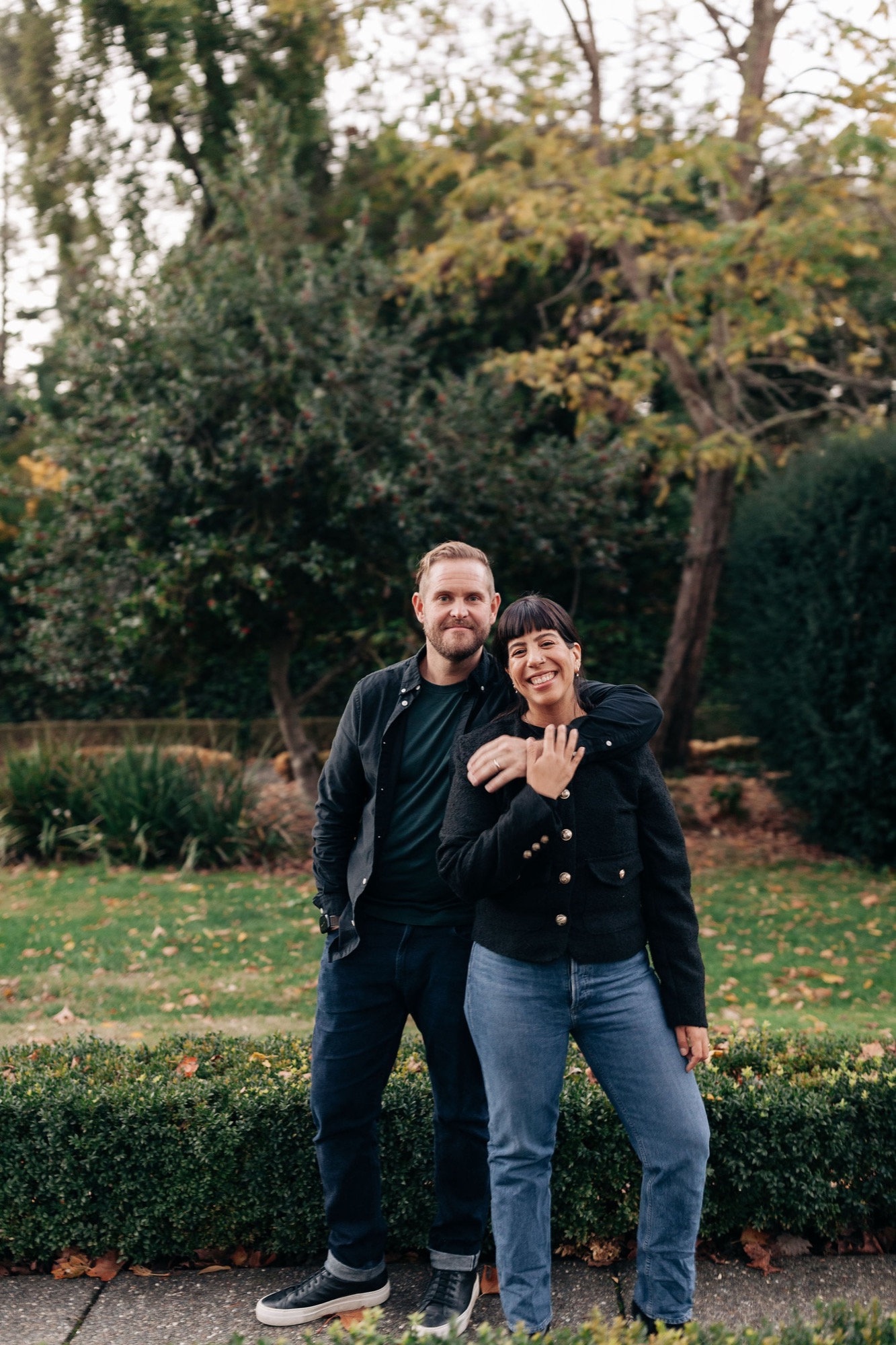 A couple stands arm-in-arm in a park wearing dark clothing, smiling warmly at the camera with autumn trees and manicured shrubs behind them
