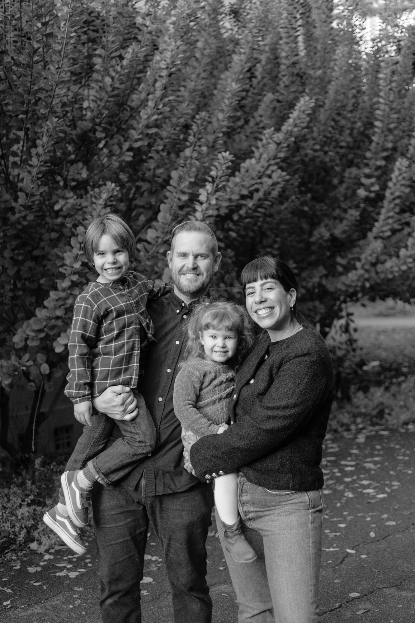 A father holds his young son while standing beside his daughter and wife against tall evergreen trees, all gazing toward the camera with gentle expressions