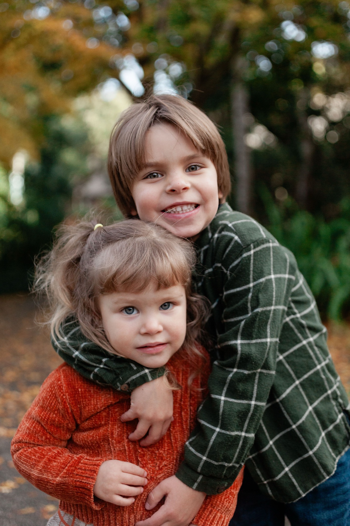 Two young siblings embrace outdoors in warm autumn light, with the older child smiling broadly while the younger looks thoughtfully toward the camera