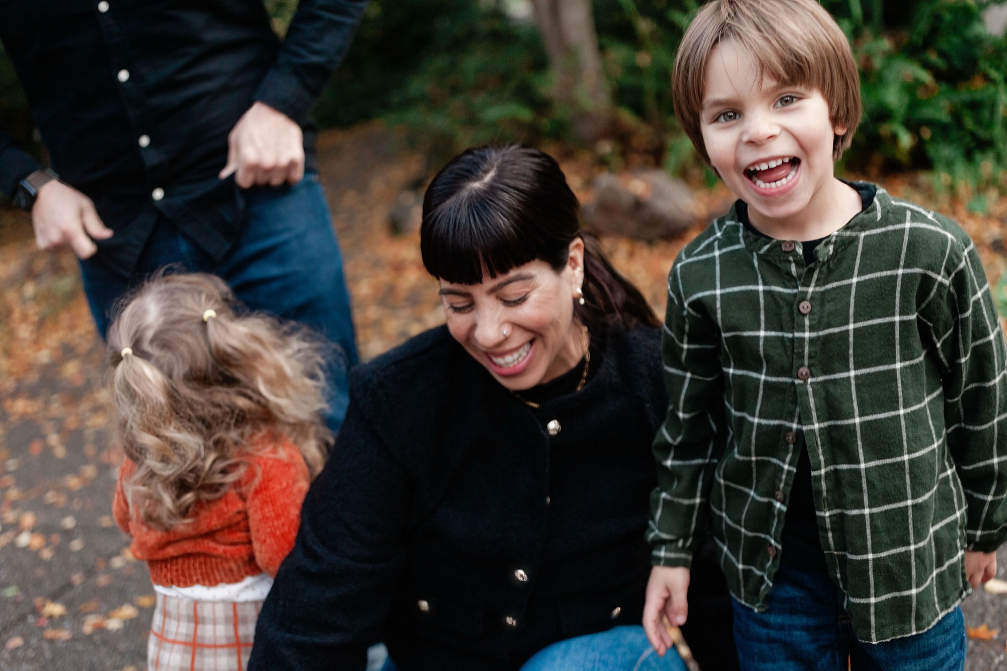 A woman crouches between two smiling children on a leaf-covered path, their genuine joy and warm connection evident in their bright expressions