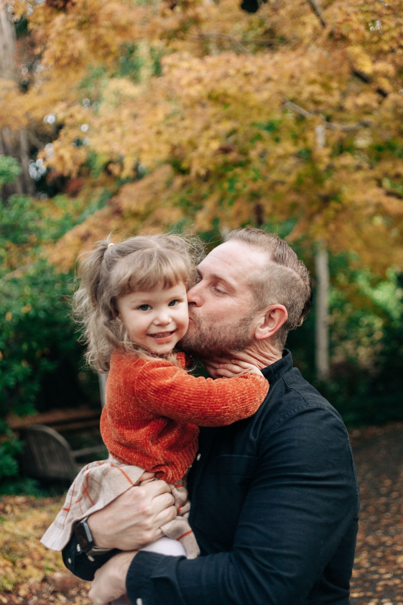 A bearded man kisses his young daughter's cheek while holding her close, golden autumn foliage glowing softly behind them