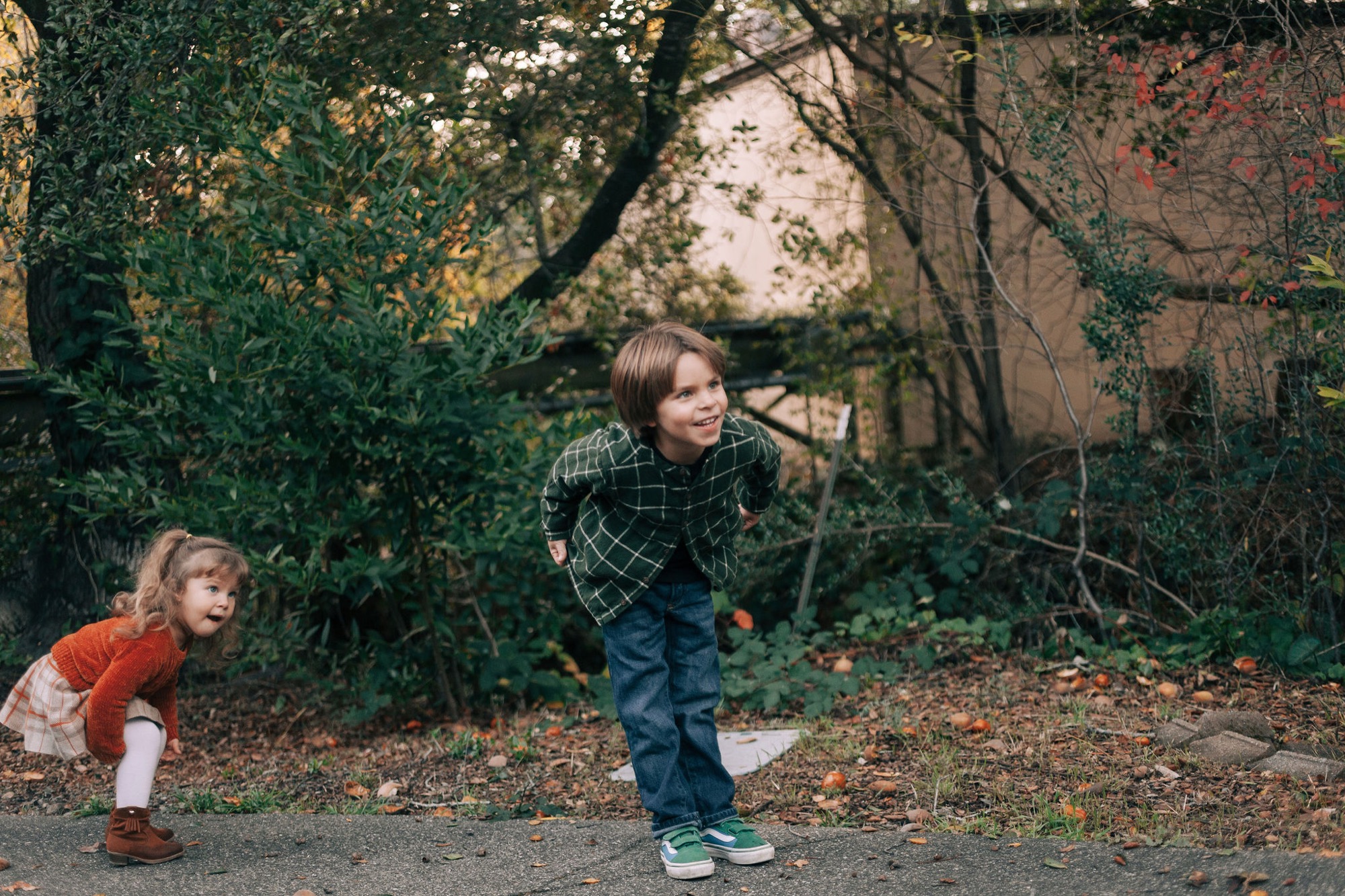 Two children play together on a leaf-covered path, their dynamic poses and engaged expressions captured in warm, dappled autumn light beneath overhanging branches