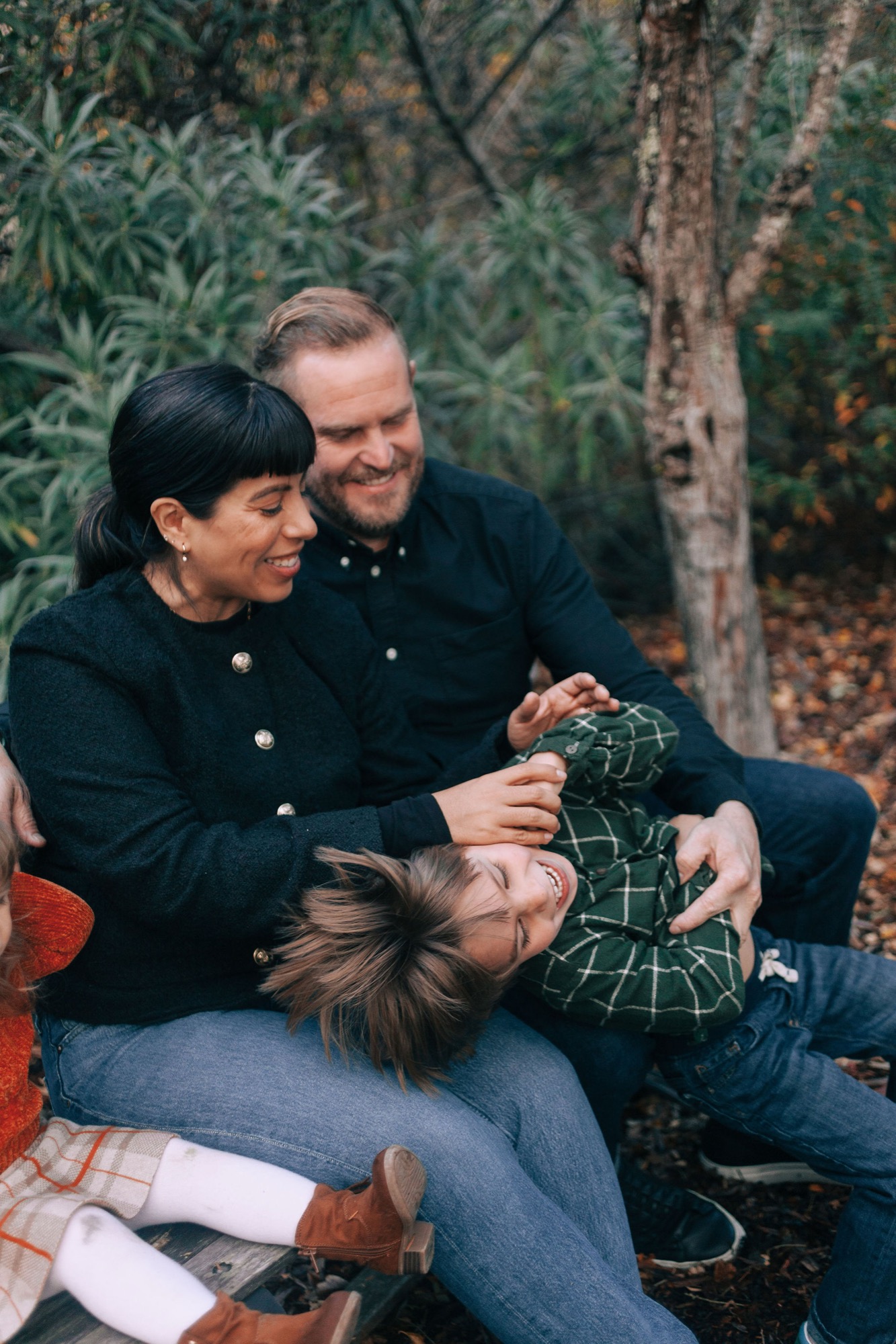 Three family members sit close together on autumn leaves, their genuine smiles and relaxed postures suggesting comfortable intimacy beneath evergreen trees