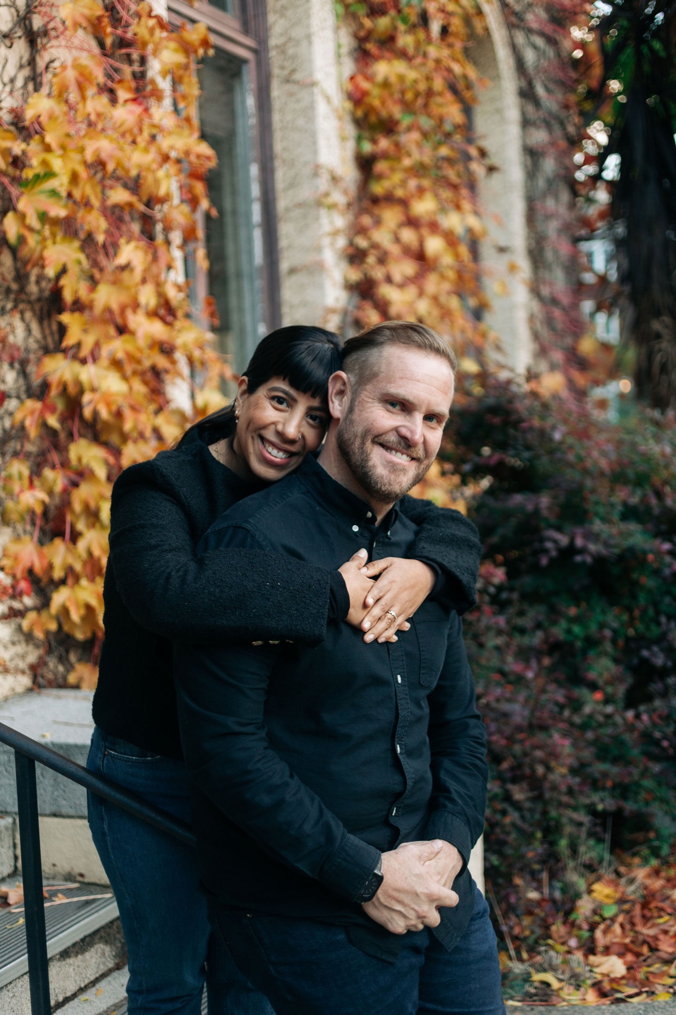 Two men embrace warmly against a building draped in golden autumn ivy, their genuine smiles and comfortable closeness radiating affection