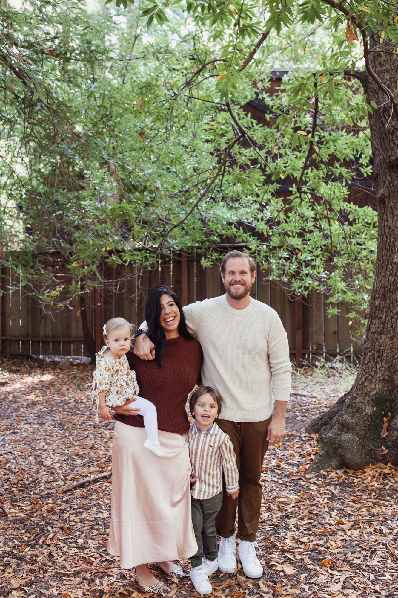 Family of four stands together on a leaf-covered forest floor beneath towering oak trees, with soft dappled light filtering through the canopy