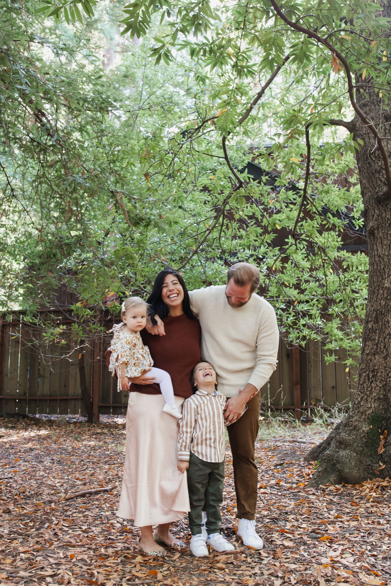 Family of four stands together on a leaf-covered forest floor beneath towering trees, mother smiling warmly while father and children lean in close