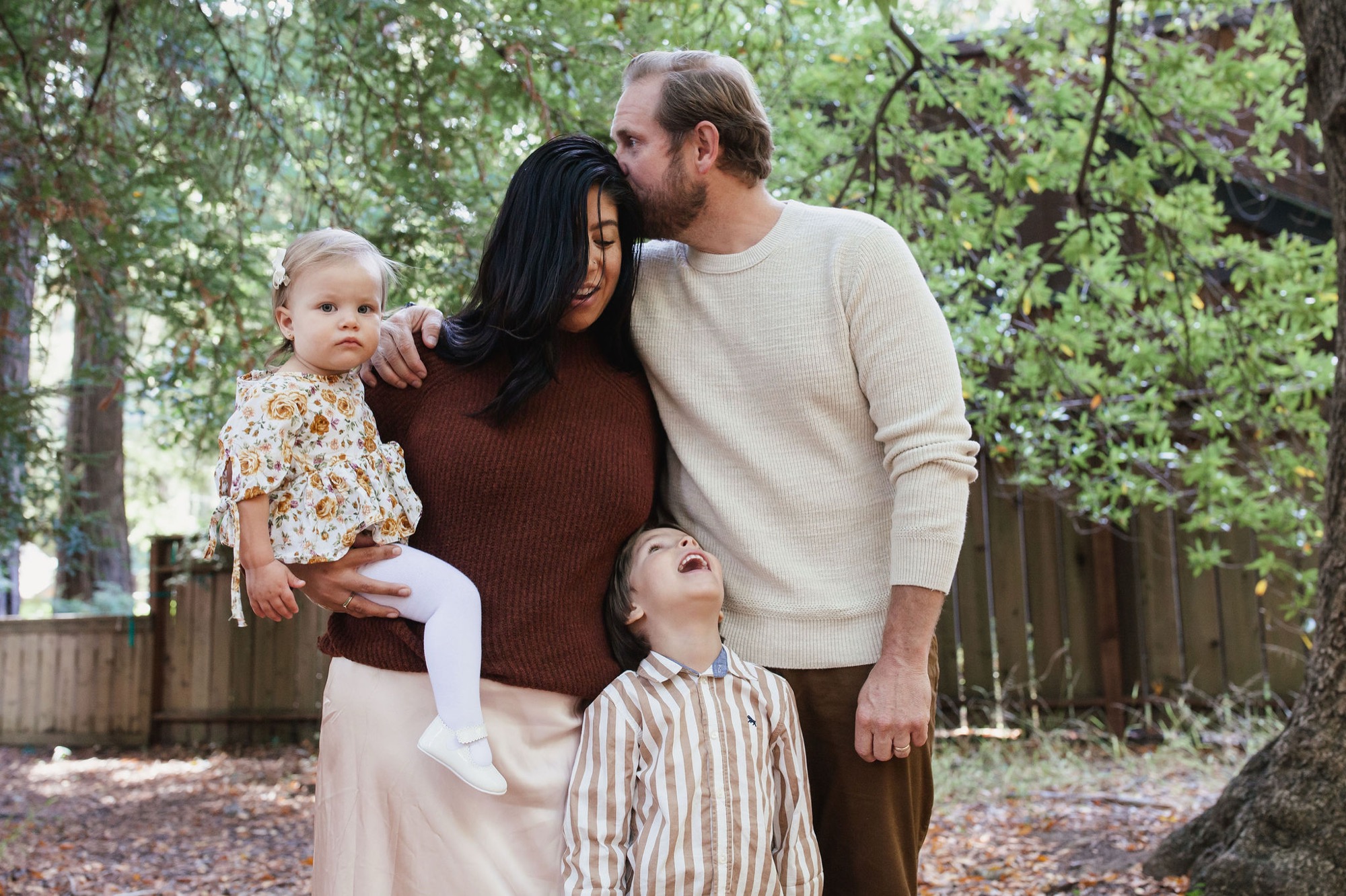 A family of four stands together among tall trees and fallen leaves, with the father kissing the mother's cheek while she holds their toddler and their young son stands close beside them