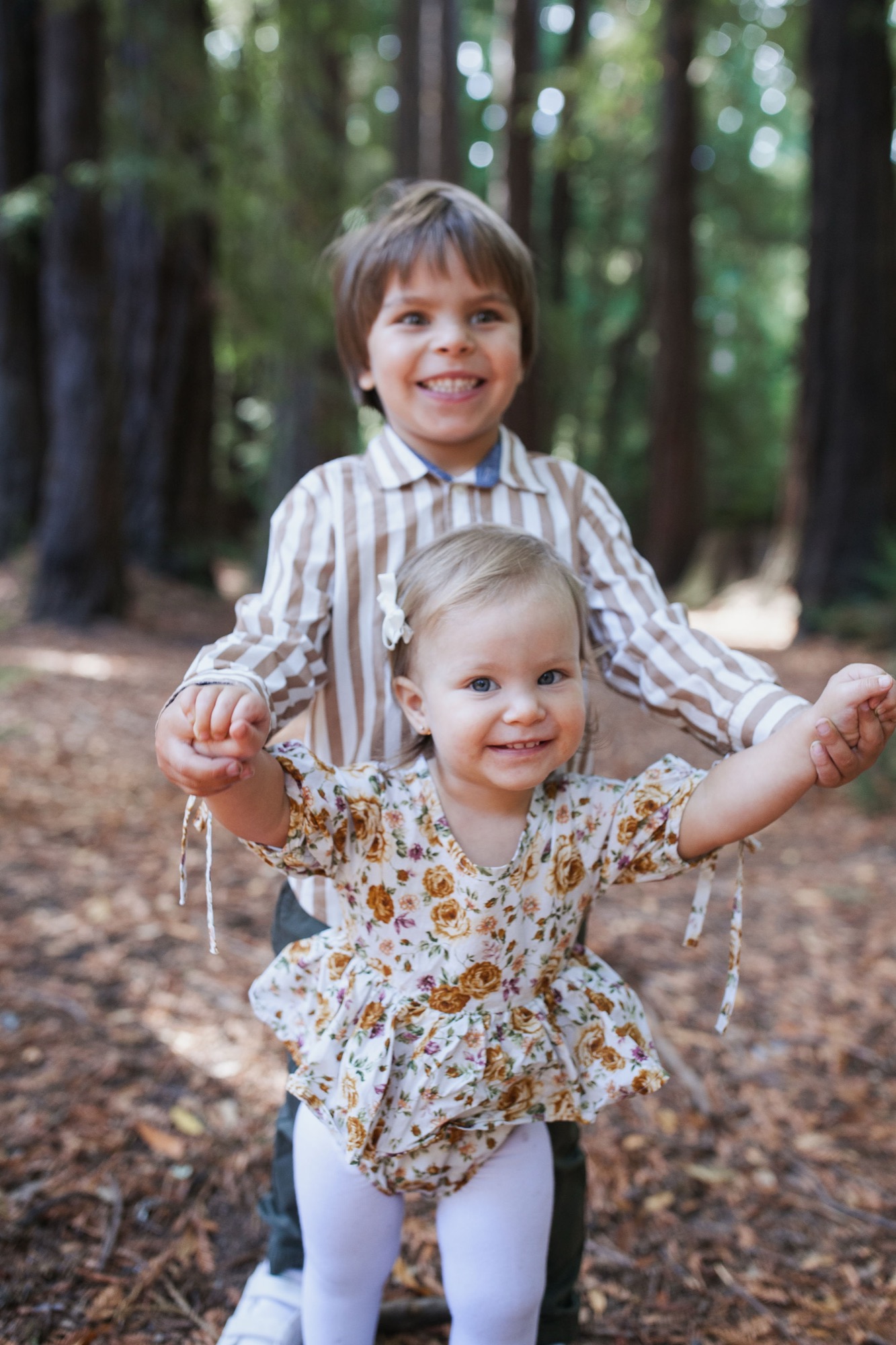 Two young children smile at the camera in a redwood forest, the older boy standing behind with arms outstretched around his younger sister in a floral dress
