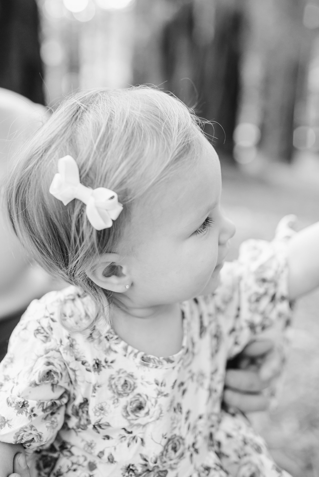 A young girl in a floral dress gazes downward, a white bow securing her blonde hair in soft profile against a blurred background