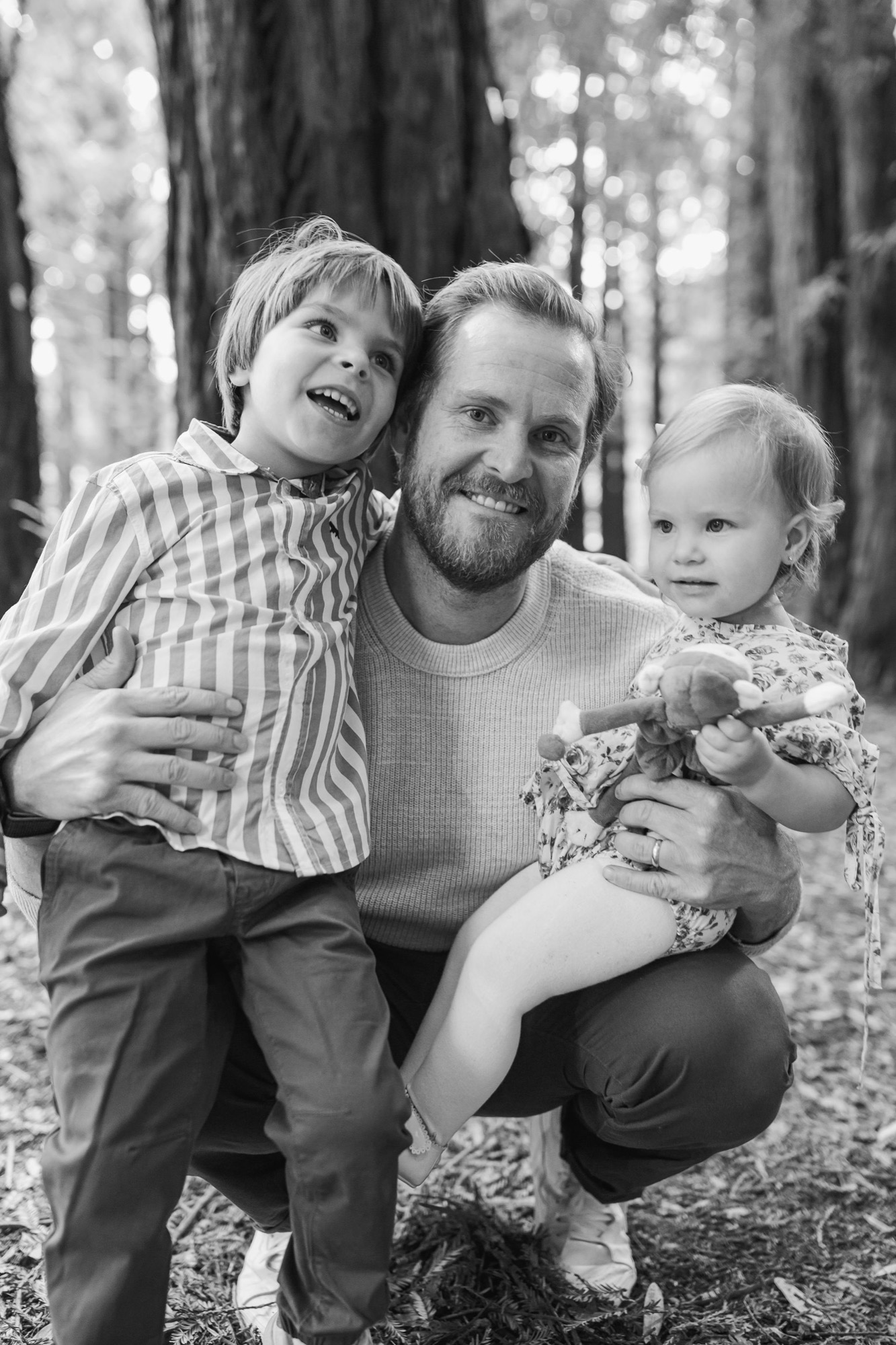 A bearded man crouches between two young children in a sunlit forest, all three smiling warmly at the camera