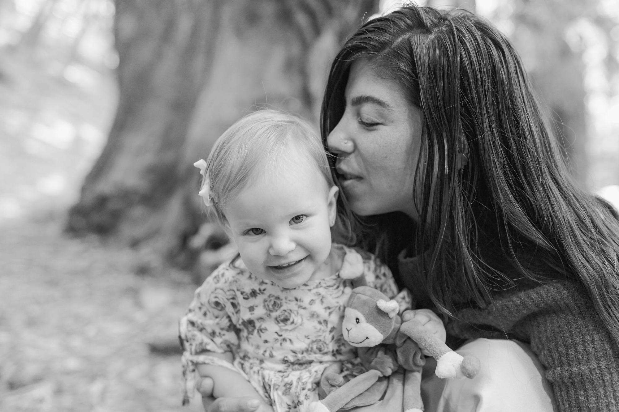 A woman leans in to kiss a blonde toddler's head while holding her close, both smiling softly in black and white outdoor light