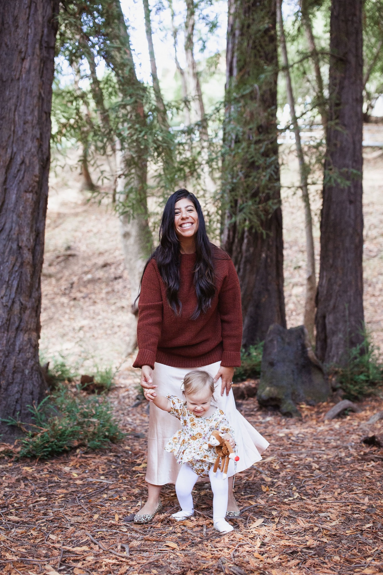Woman in burgundy sweater and white ripped jeans smiles among towering redwoods on a leaf-covered forest floor