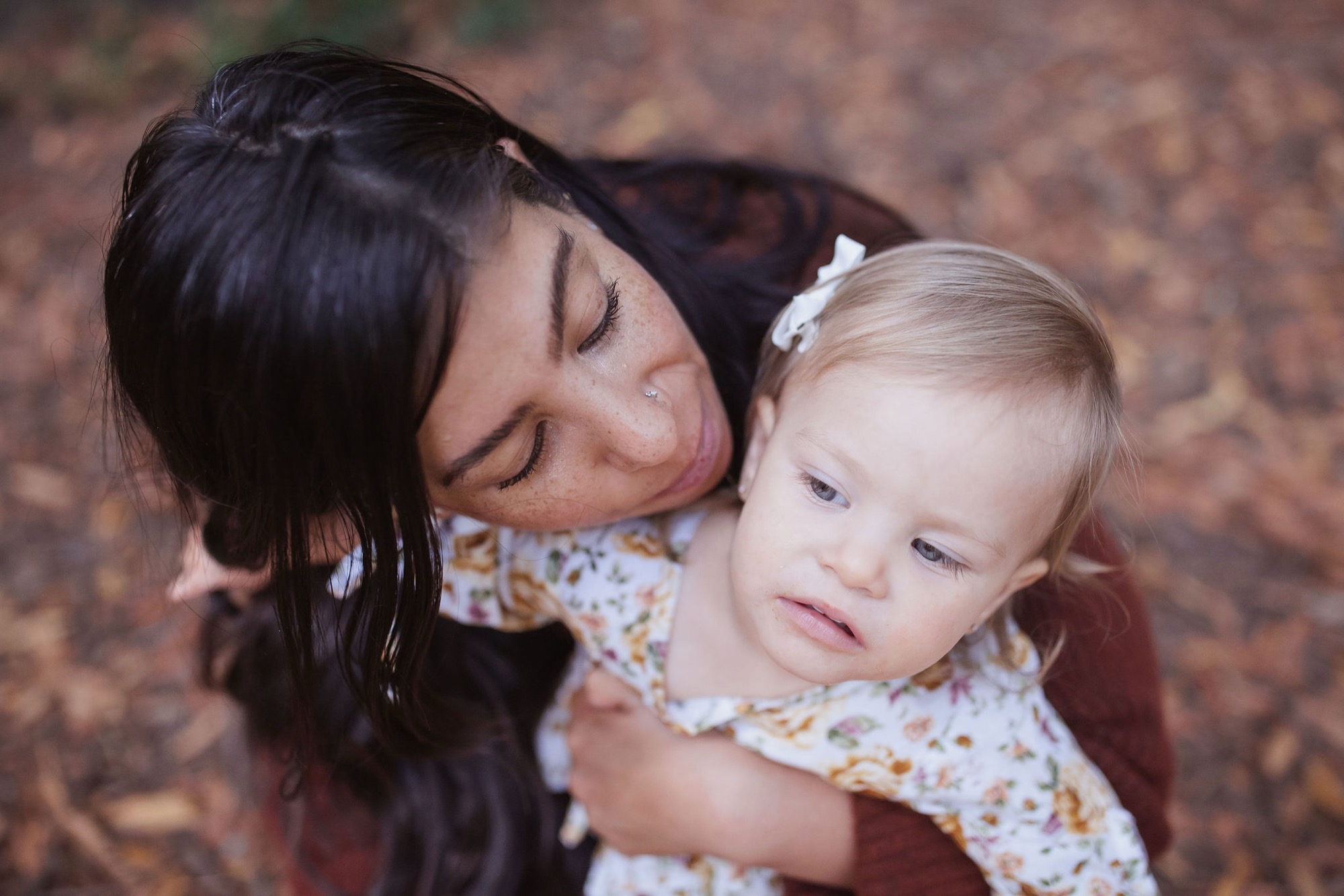 A mother with dark hair closes her eyes while tenderly holding her blonde toddler against her shoulder in warm afternoon light
