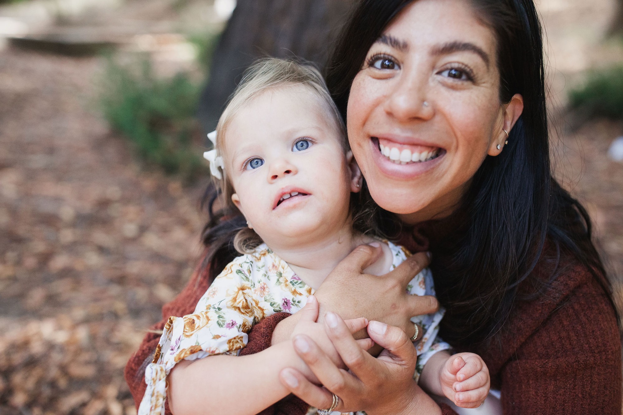 A woman in a burgundy cardigan holds a blonde toddler who gazes upward with wonder while she beams at the camera on a dusty outdoor path