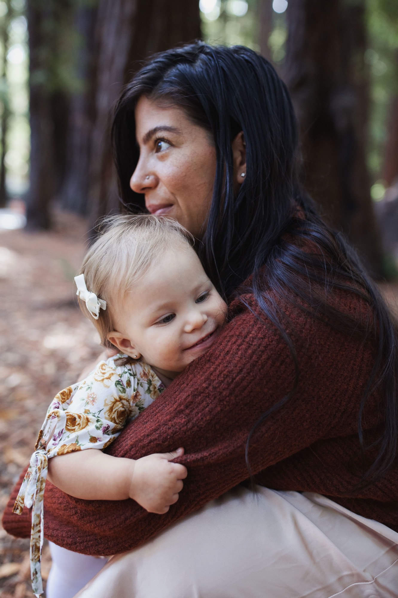 A woman holds her toddler in a forest setting, both smiling warmly at the camera with dappled sunlight filtering through tall trees