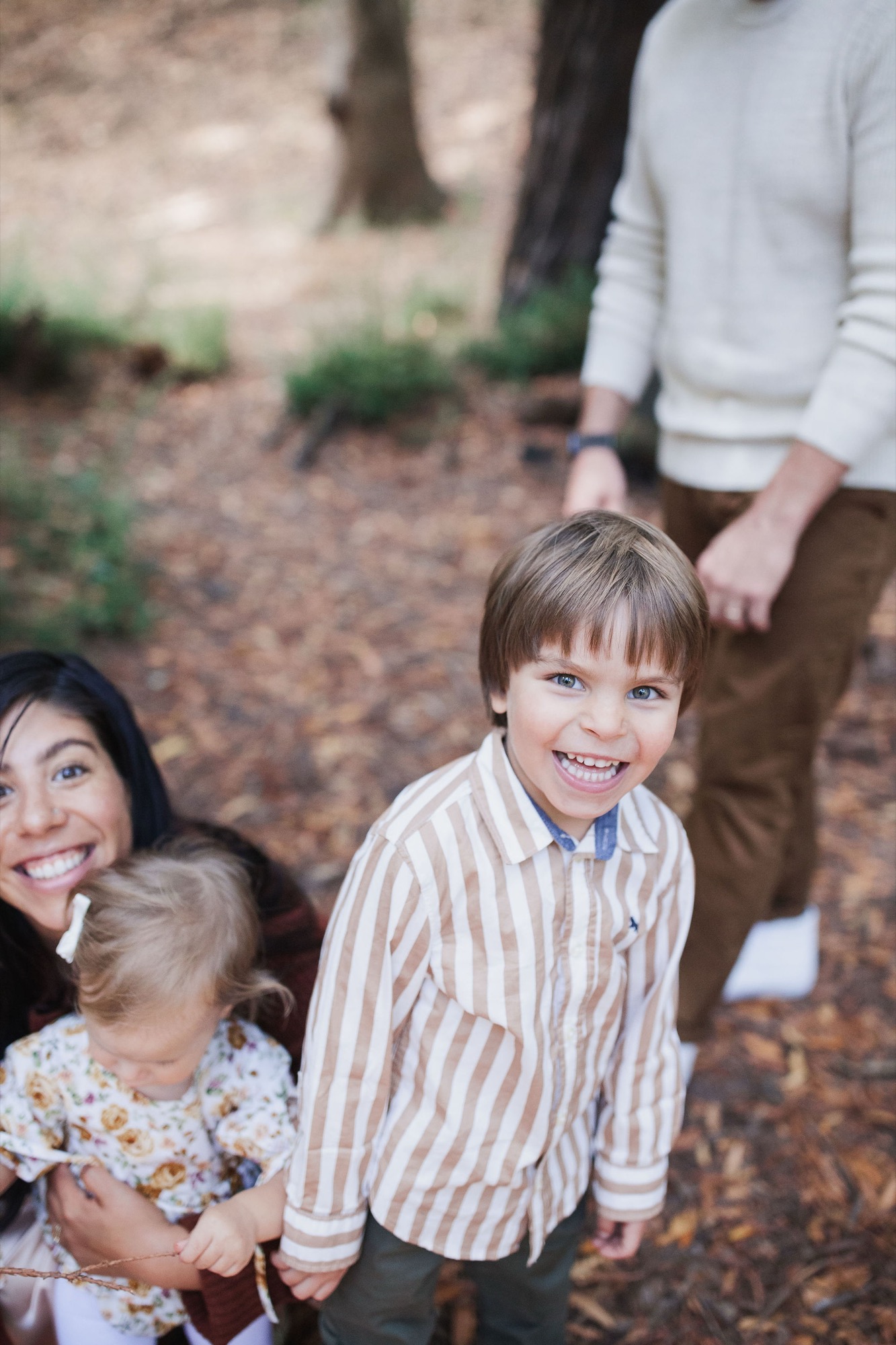Two smiling children in striped and floral shirts stand on a pine needle path while adults stand behind them in earth-toned clothing
